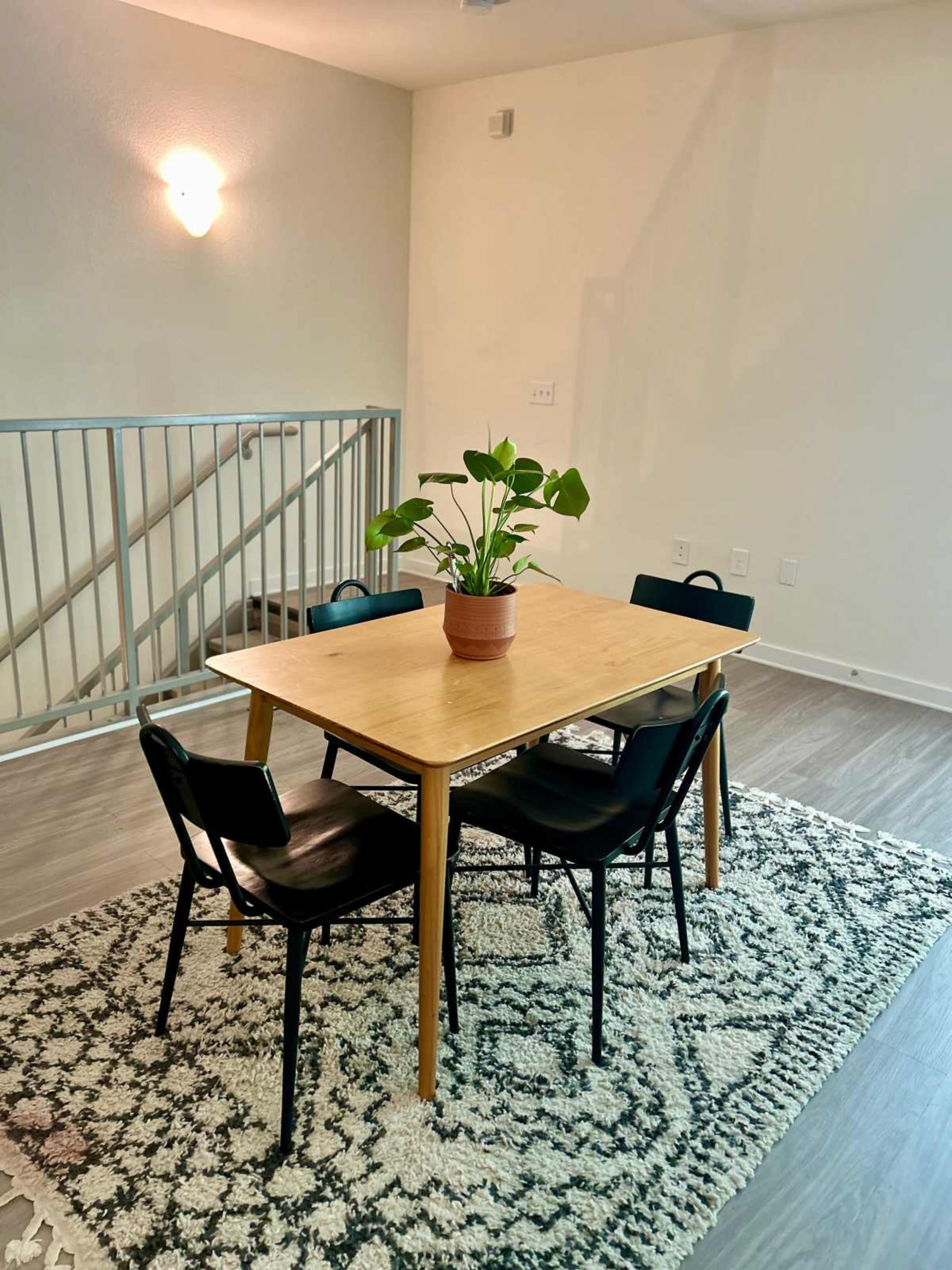 A wooden table surrounded by four black chairs sits on a patterned rug in a brightly lit room with a potted plant in the center.