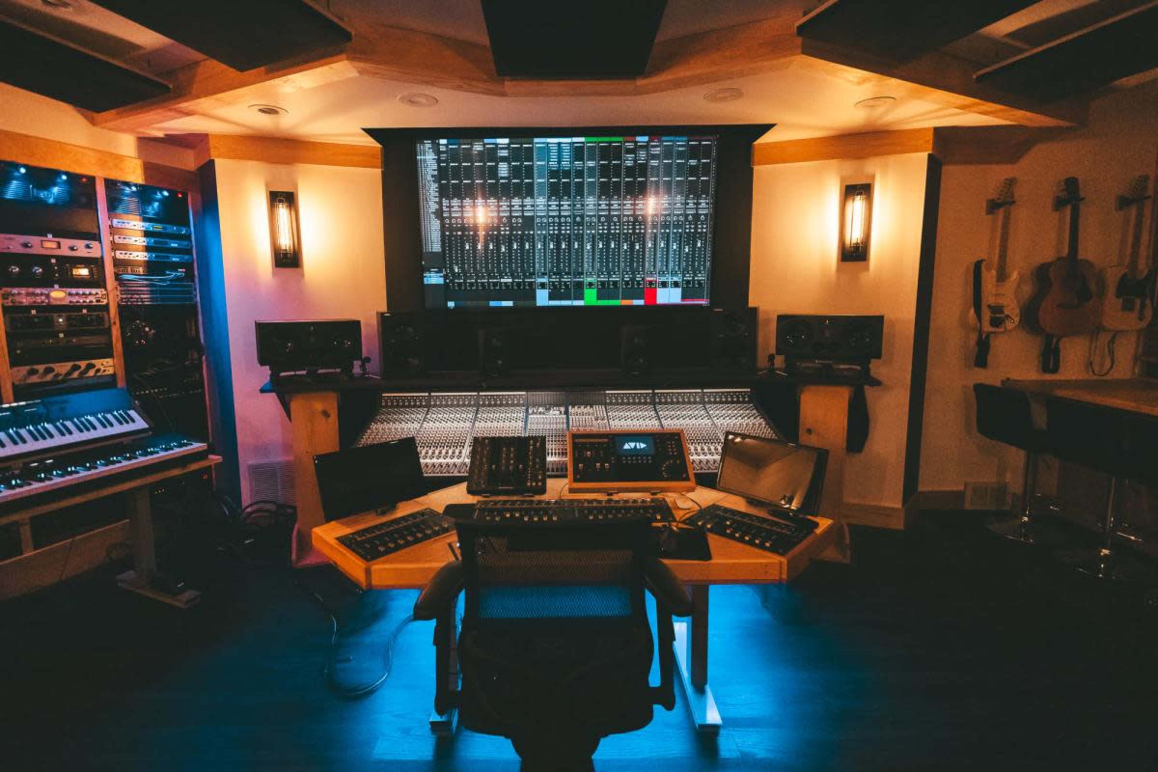 The image shows a well-equipped audio recording studio with a large mixing console, computer monitors, and various musical instruments on the walls.