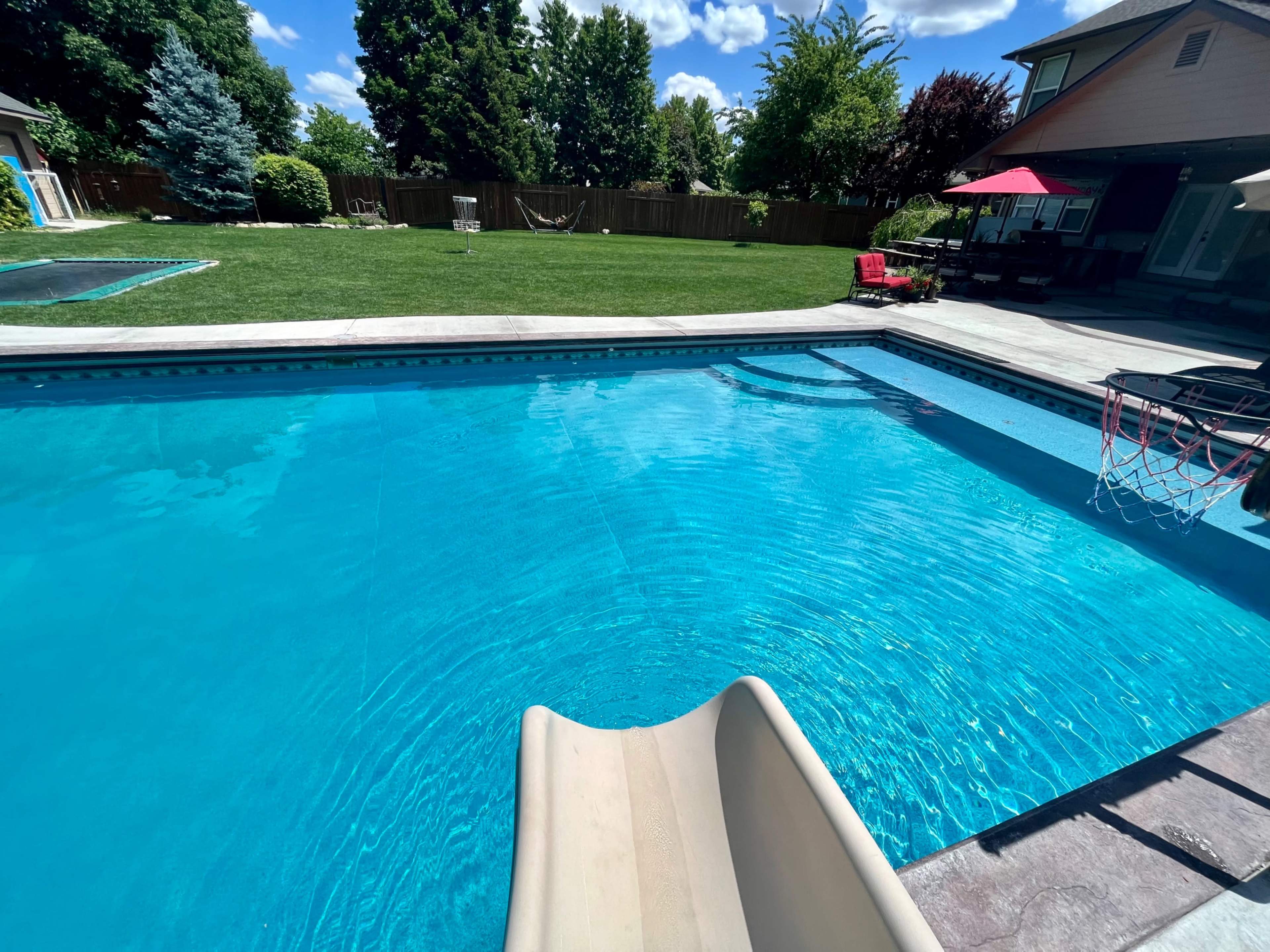 The image shows a clear swimming pool with a slide, surrounded by green grass and trees, and a patio area with seating and an umbrella.