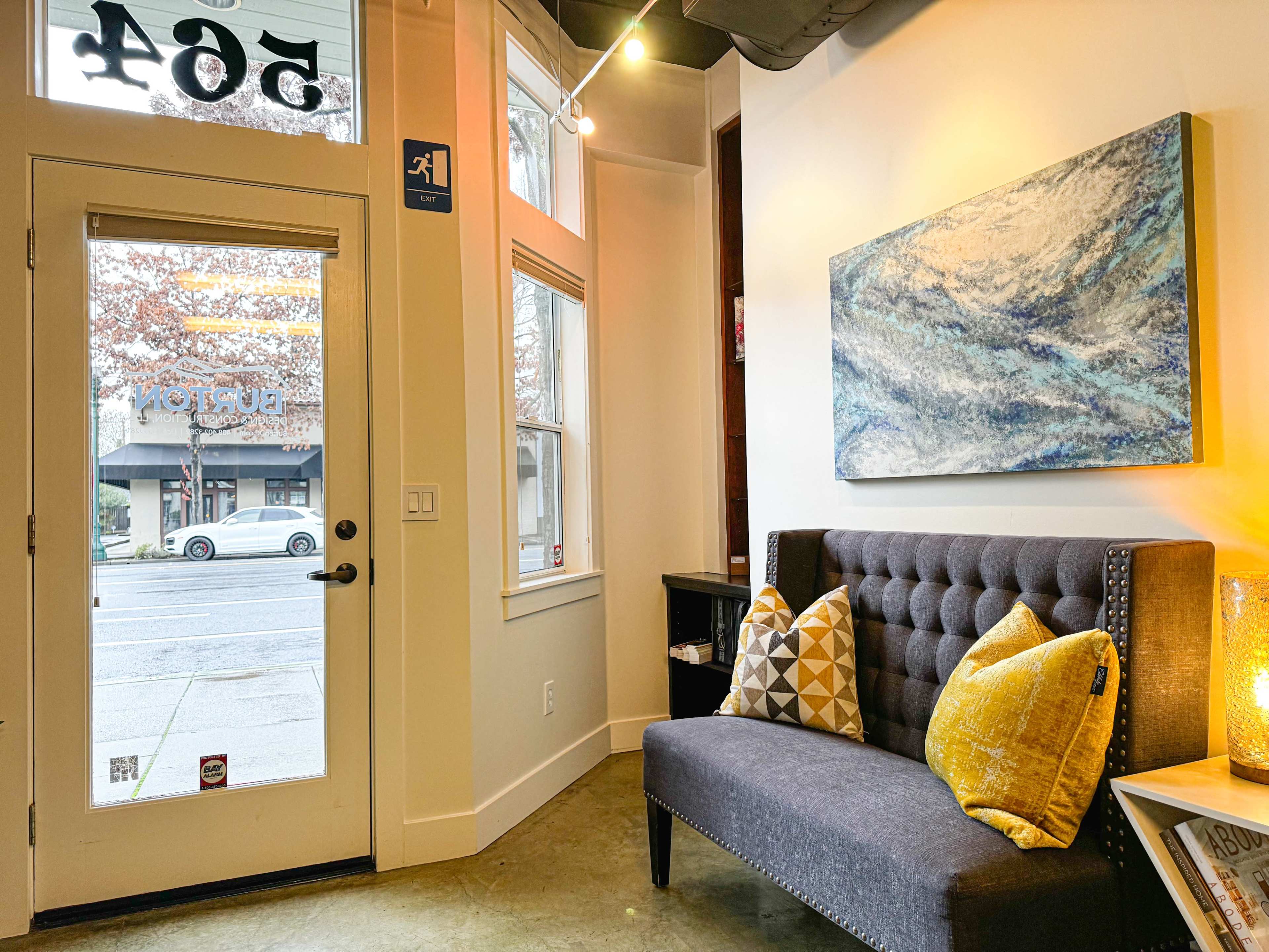 A cozy seating area with a gray upholstered couch adorned with yellow and white patterned pillows, positioned near a large window and a glass door that opens to the street outside.