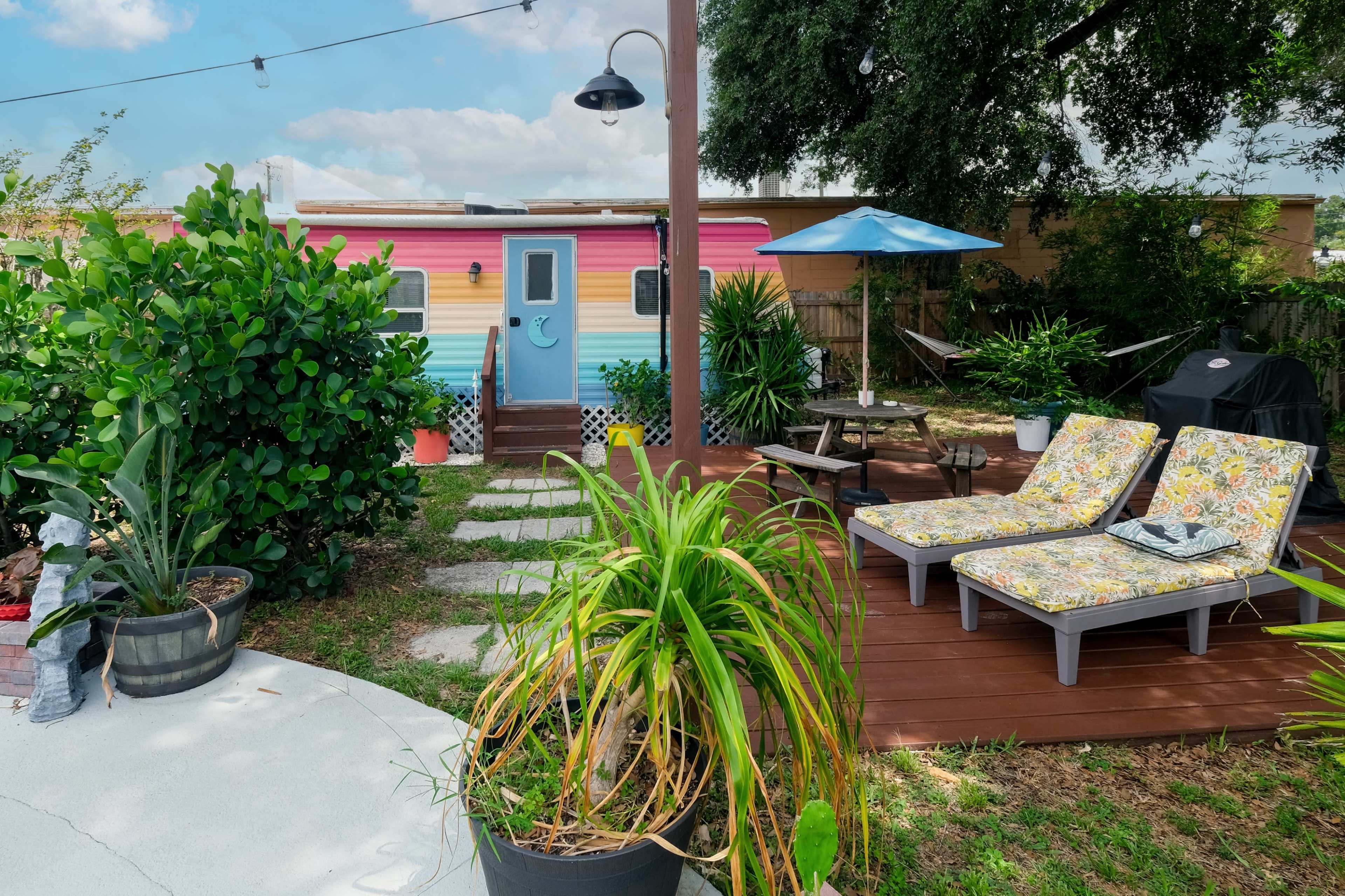 The image shows a vibrant backyard with colorful lounge chairs, a small blue umbrella, and a trailer in the background, surrounded by greenery and stone pathways.