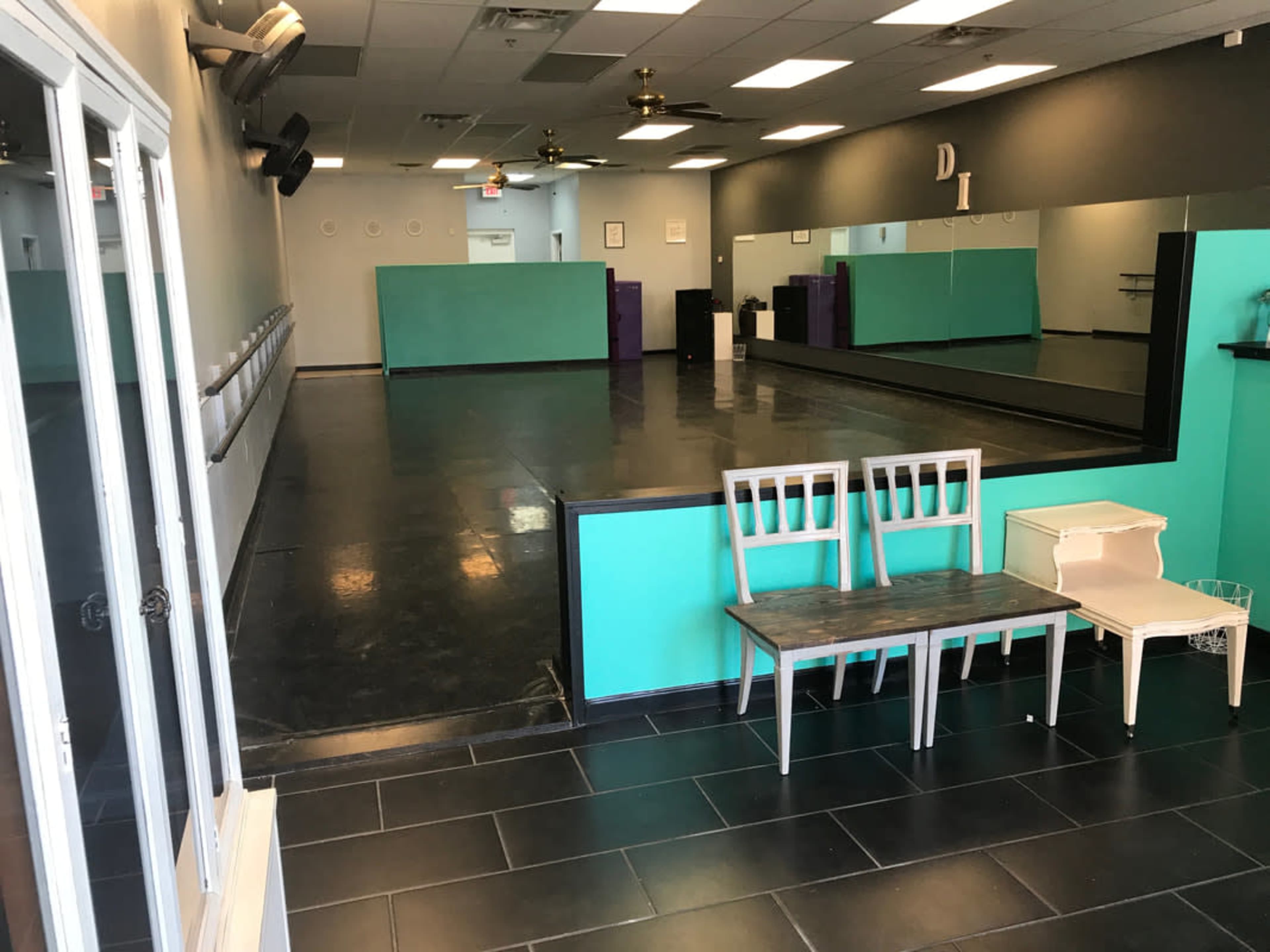 The image shows a spacious dance studio with a mirrored wall and a black floor, featuring teal and purple accents, along with two chairs and a small table.
