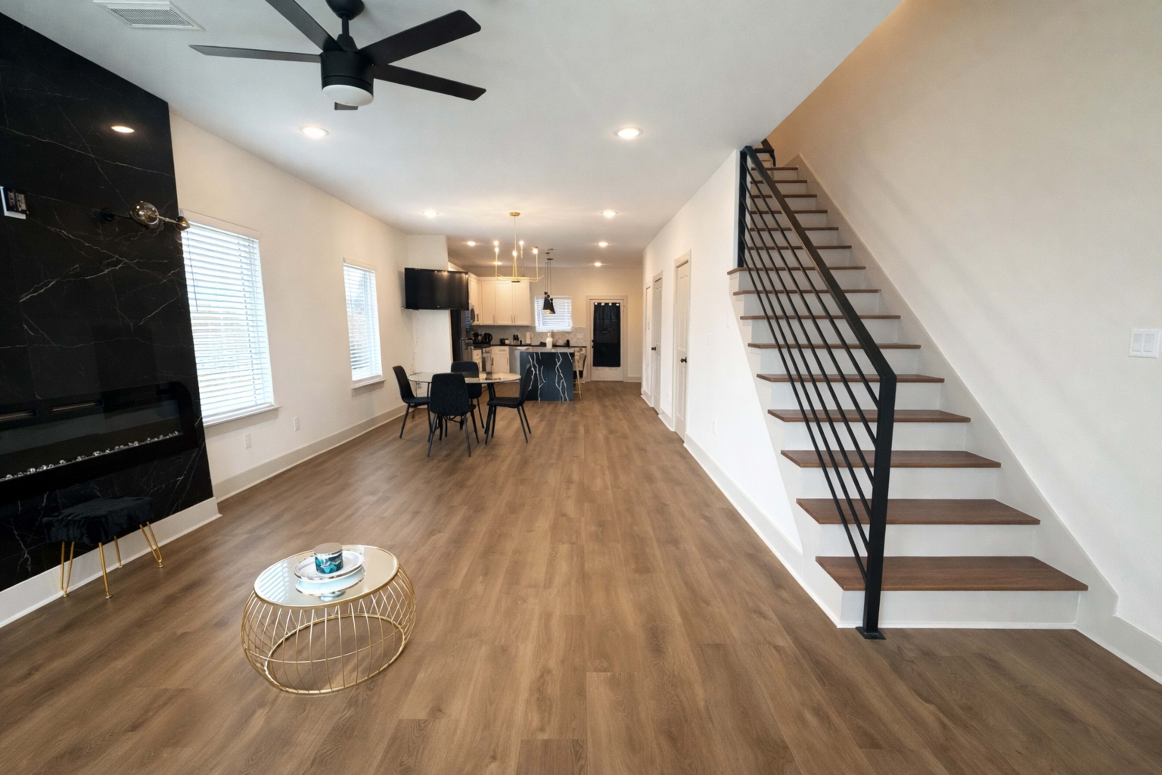 The image depicts a modern open-concept living area with a staircase on the right, a dining space, and a kitchen visible in the background.
