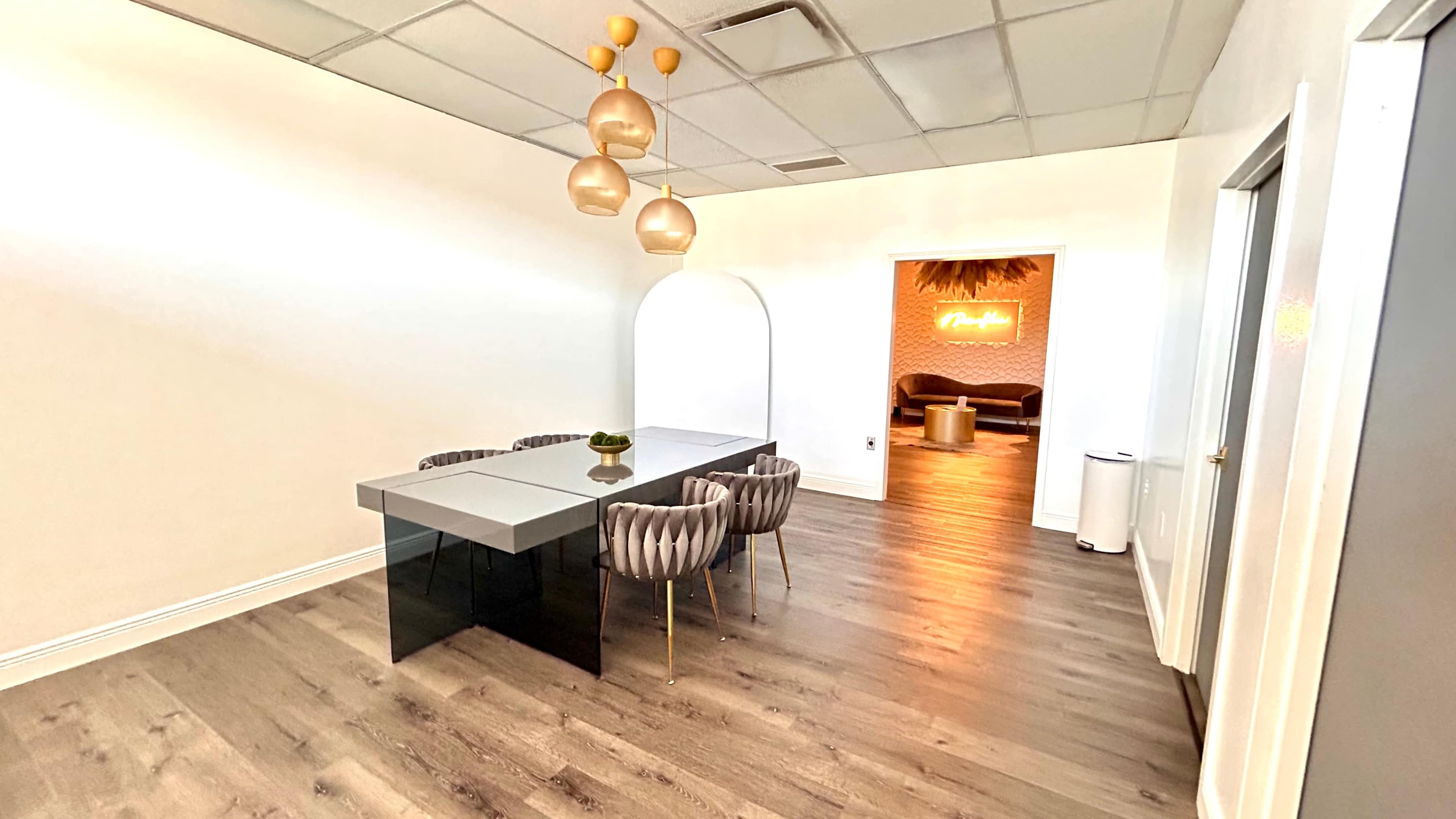 A modern conference room features a sleek glass table surrounded by tufted chairs, with a doorway leading to a lounge area illuminated by neon signage.