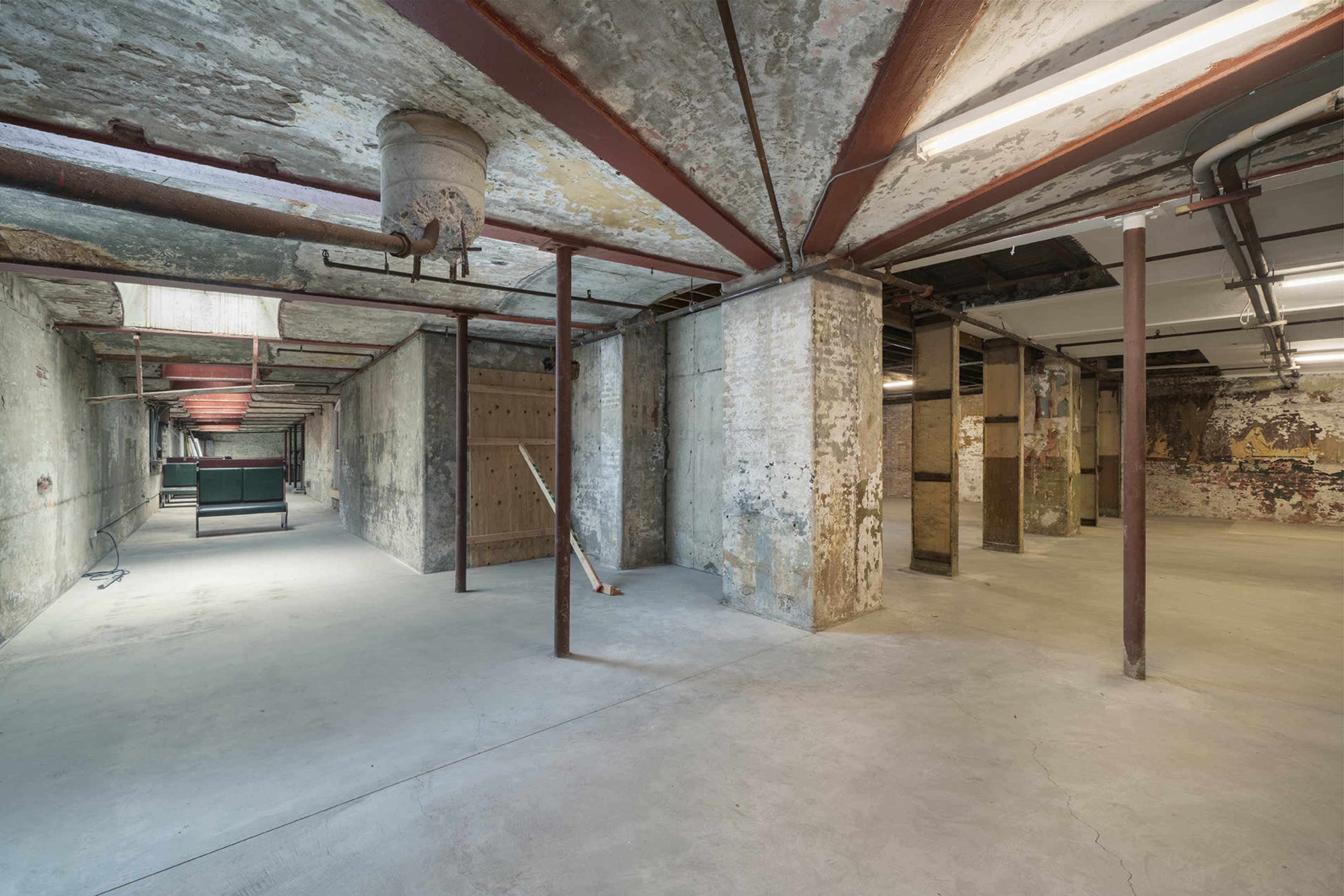 A large, empty basement space with exposed concrete walls, metal pillars, and a few overhead lights illuminating the area.