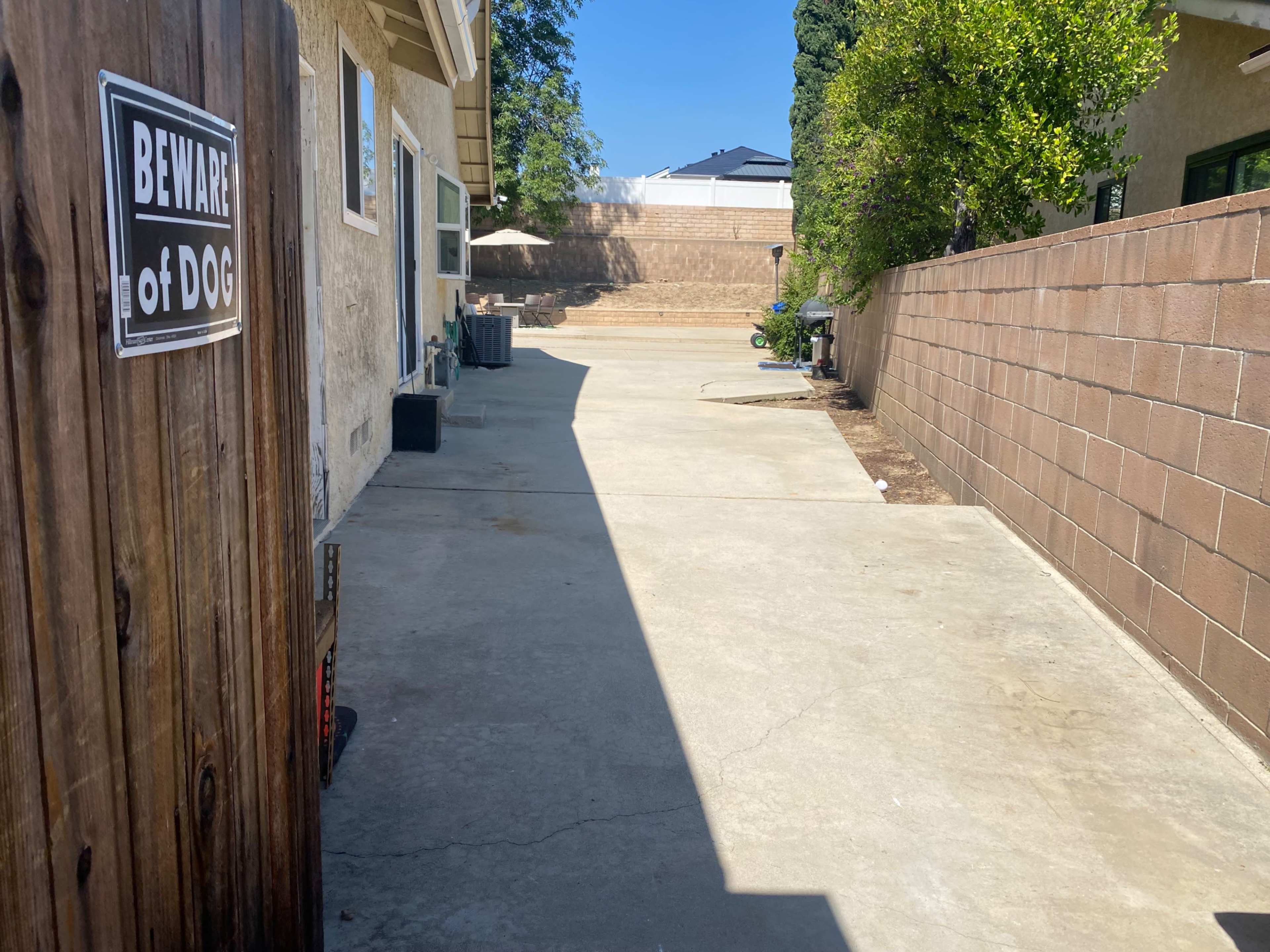A concrete pathway runs between two buildings, bordered by a wooden fence and a warning sign about a dog.