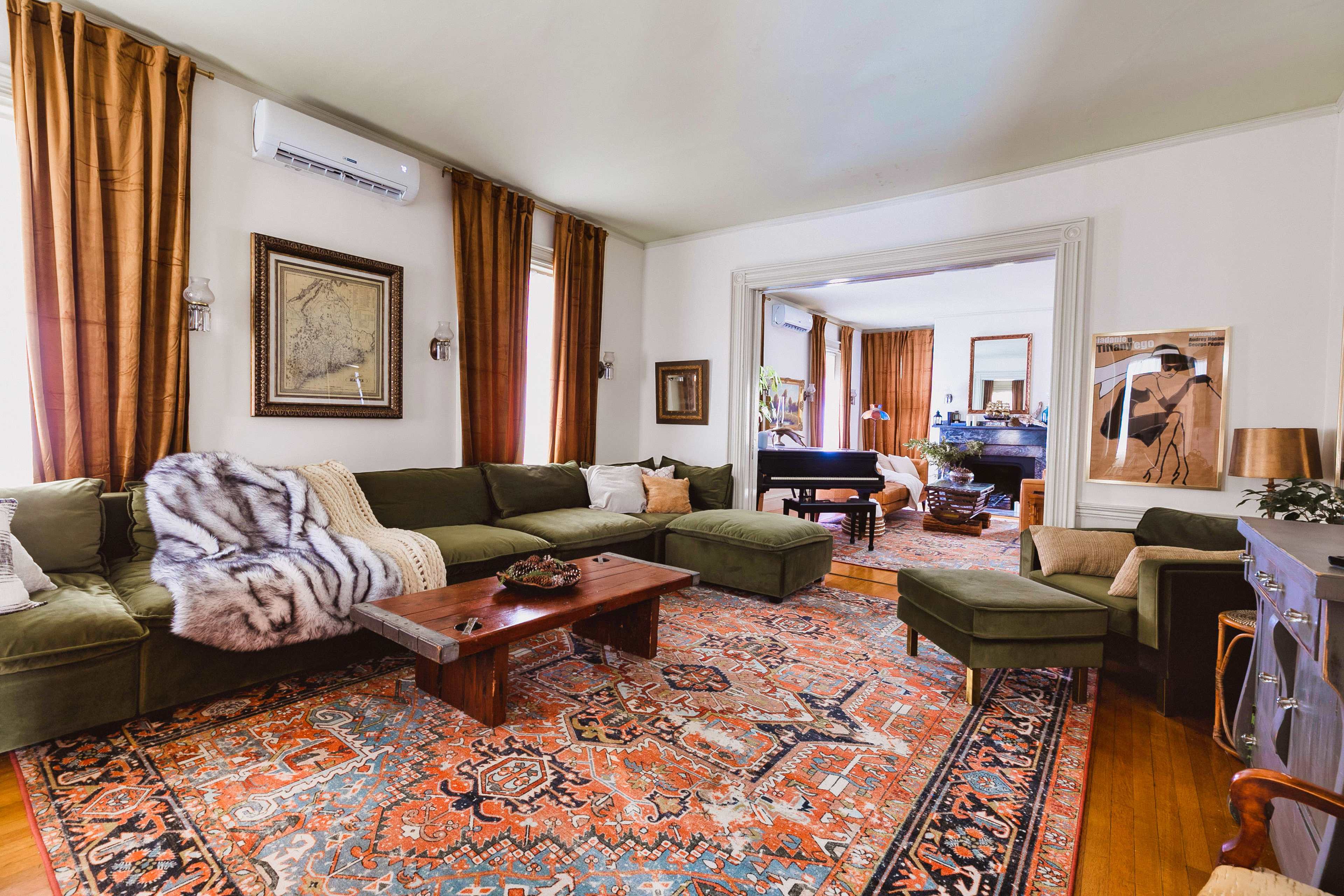 The living room features a large green sectional sofa, a wooden coffee table, an area rug with intricate patterns, and decorative curtains framing the windows.