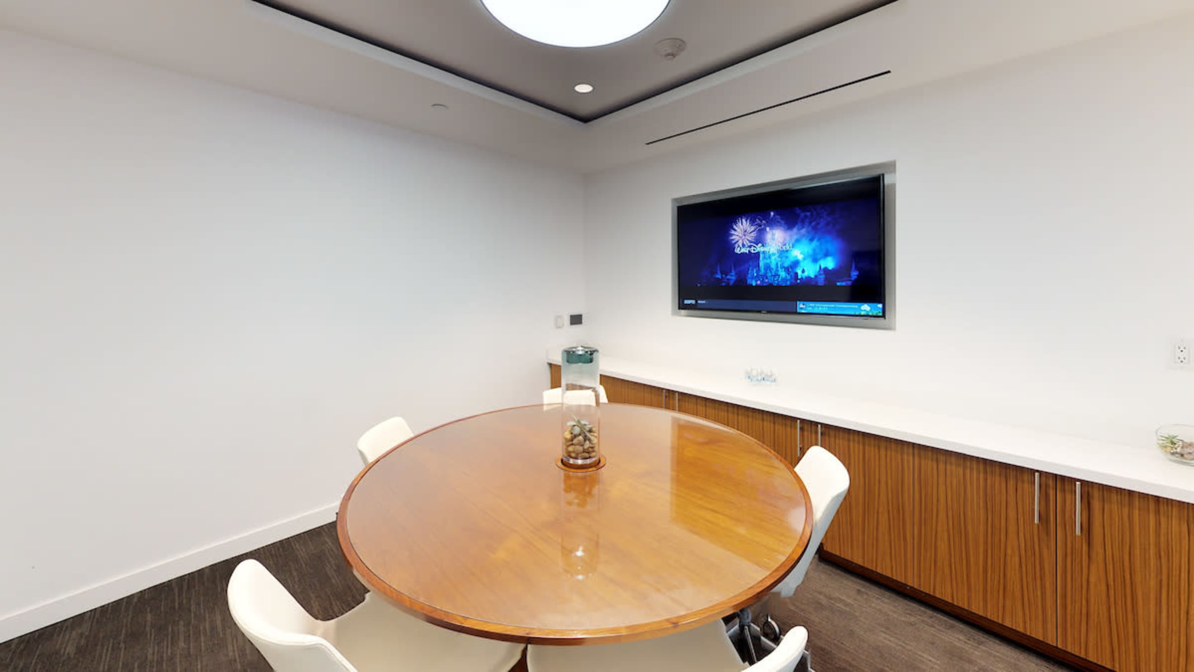 A round wooden table with white chairs is positioned in a well-lit room featuring a television mounted on the wall.