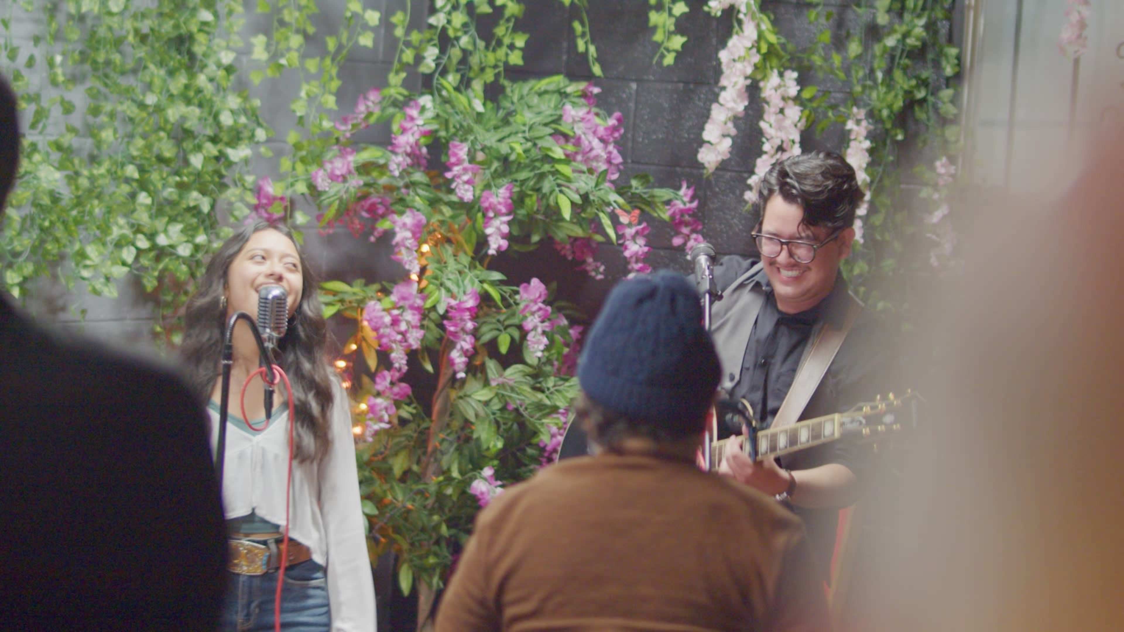 A man plays guitar while a woman sings into a microphone, both performing in front of a backdrop of flowers and greenery.