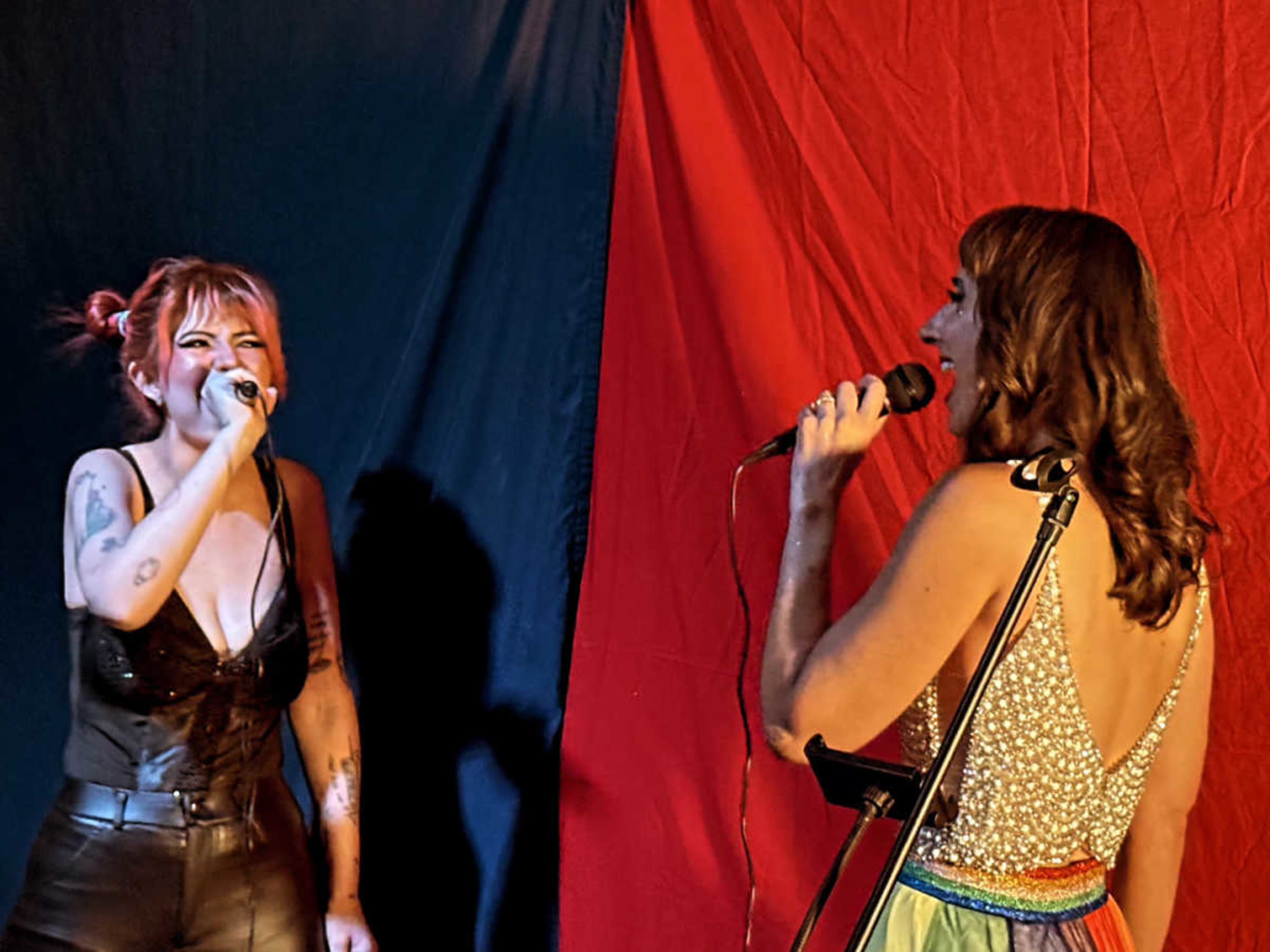 Two performers sing into microphones on a stage with a blue and red backdrop.