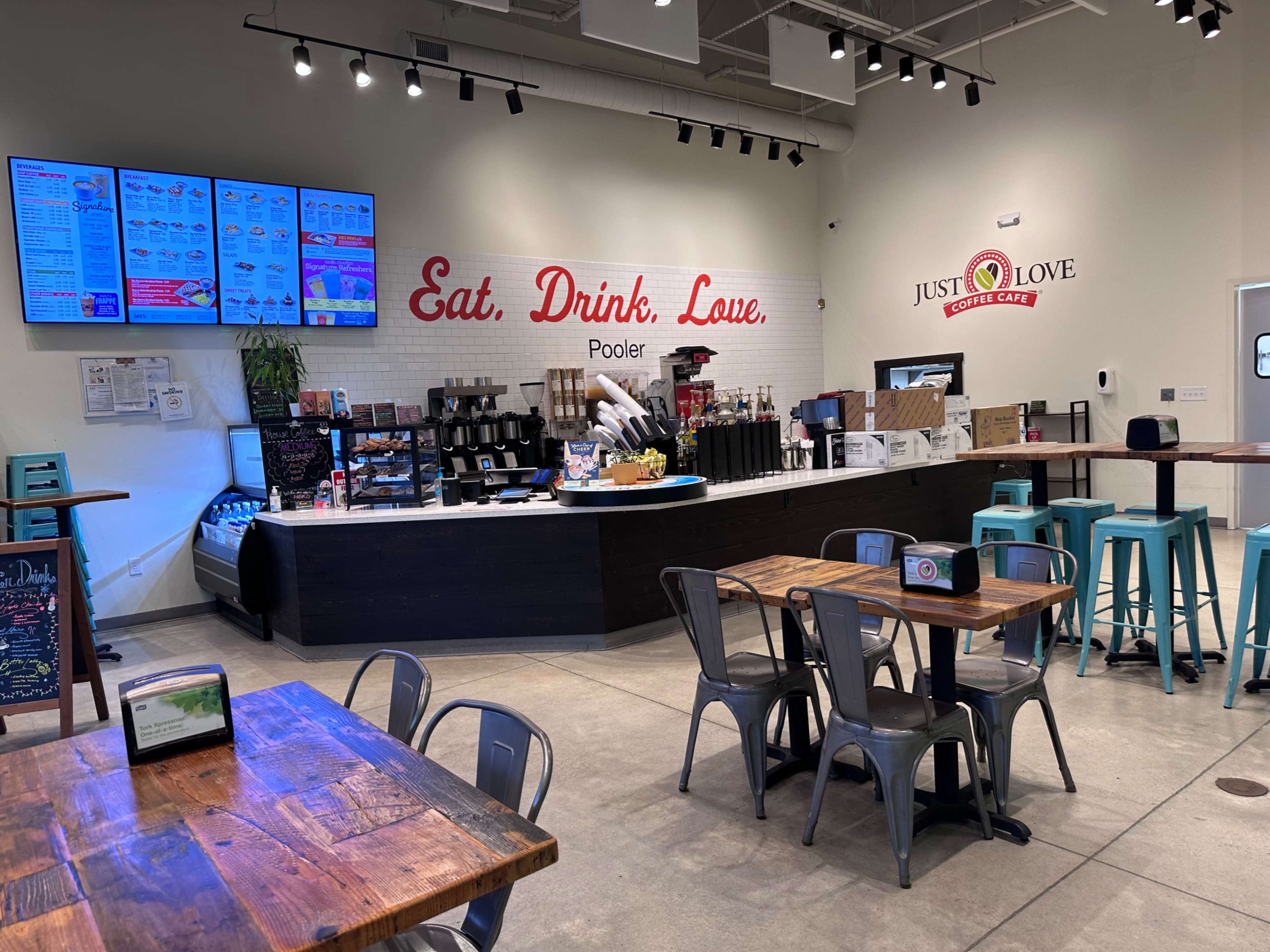 A modern cafe interior with a counter displaying menus and various food items, and seating arrangements consisting of wooden tables and metal chairs.