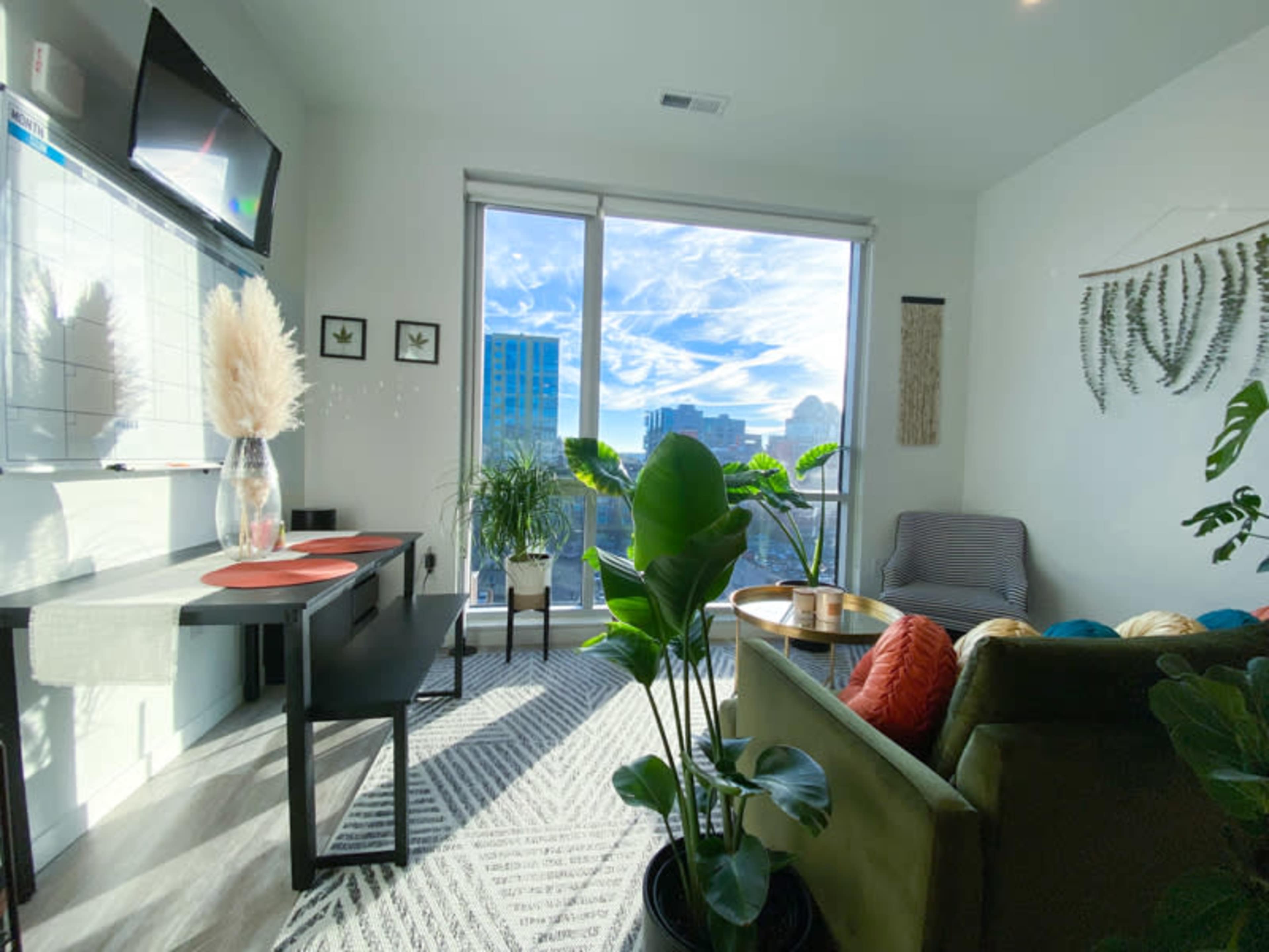 A modern living room features a green sofa, a dining table with chairs, and large windows offering a city skyline view.