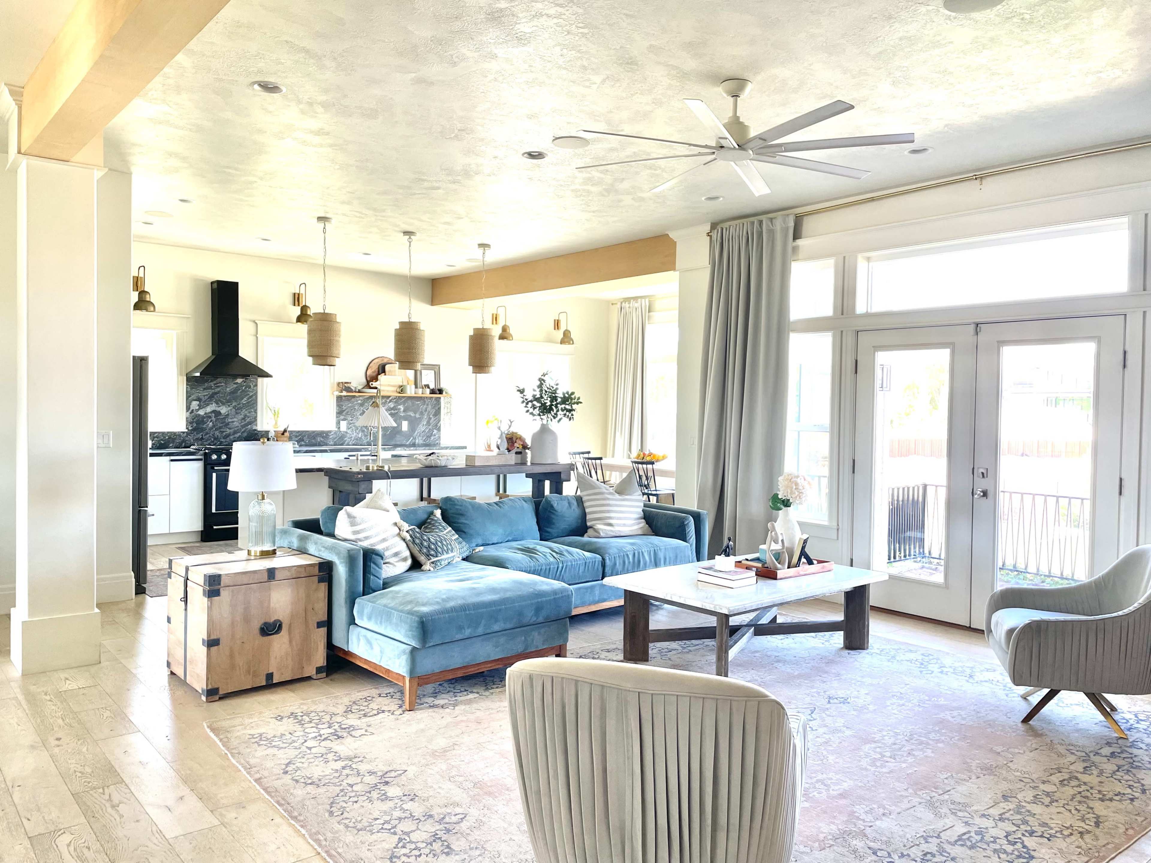 A bright, airy living room featuring a large blue sectional sofa, a wooden coffee table, and large windows that let in natural light.