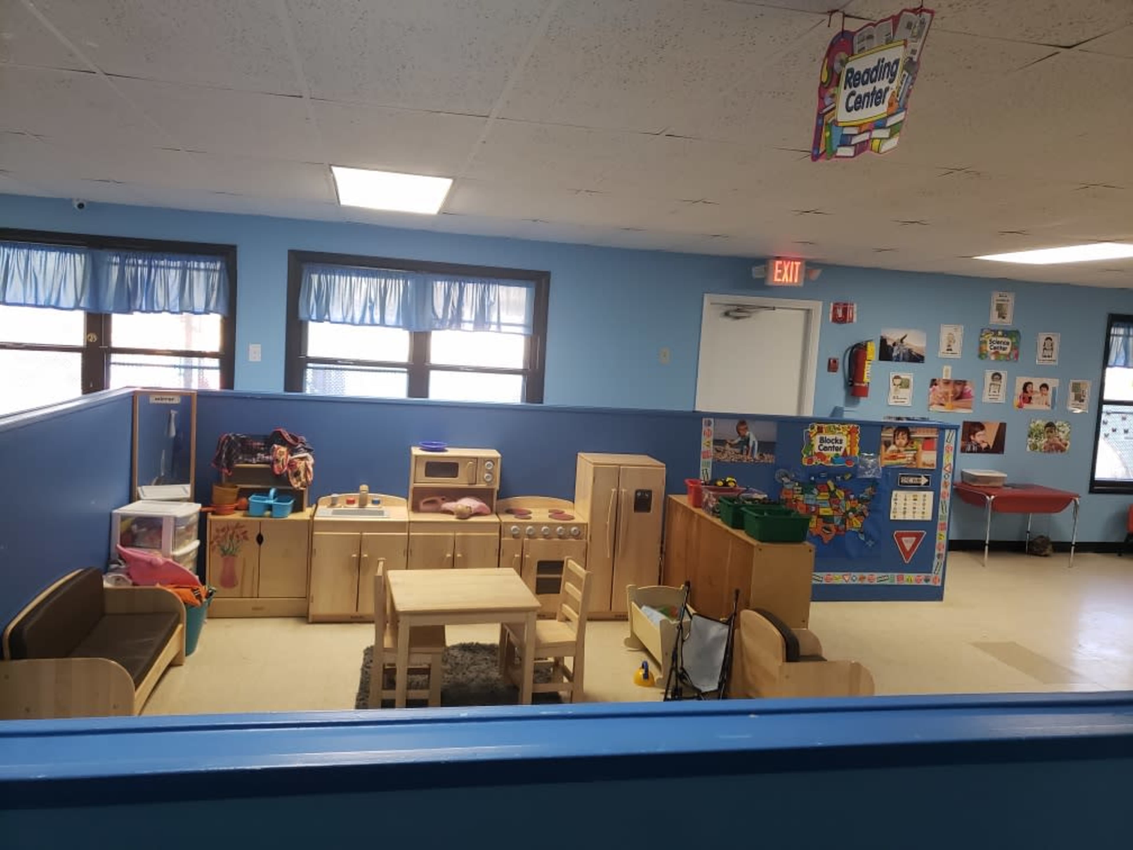A brightly lit classroom contains various educational toys, a play kitchen, and a reading area with children's artwork on the walls.