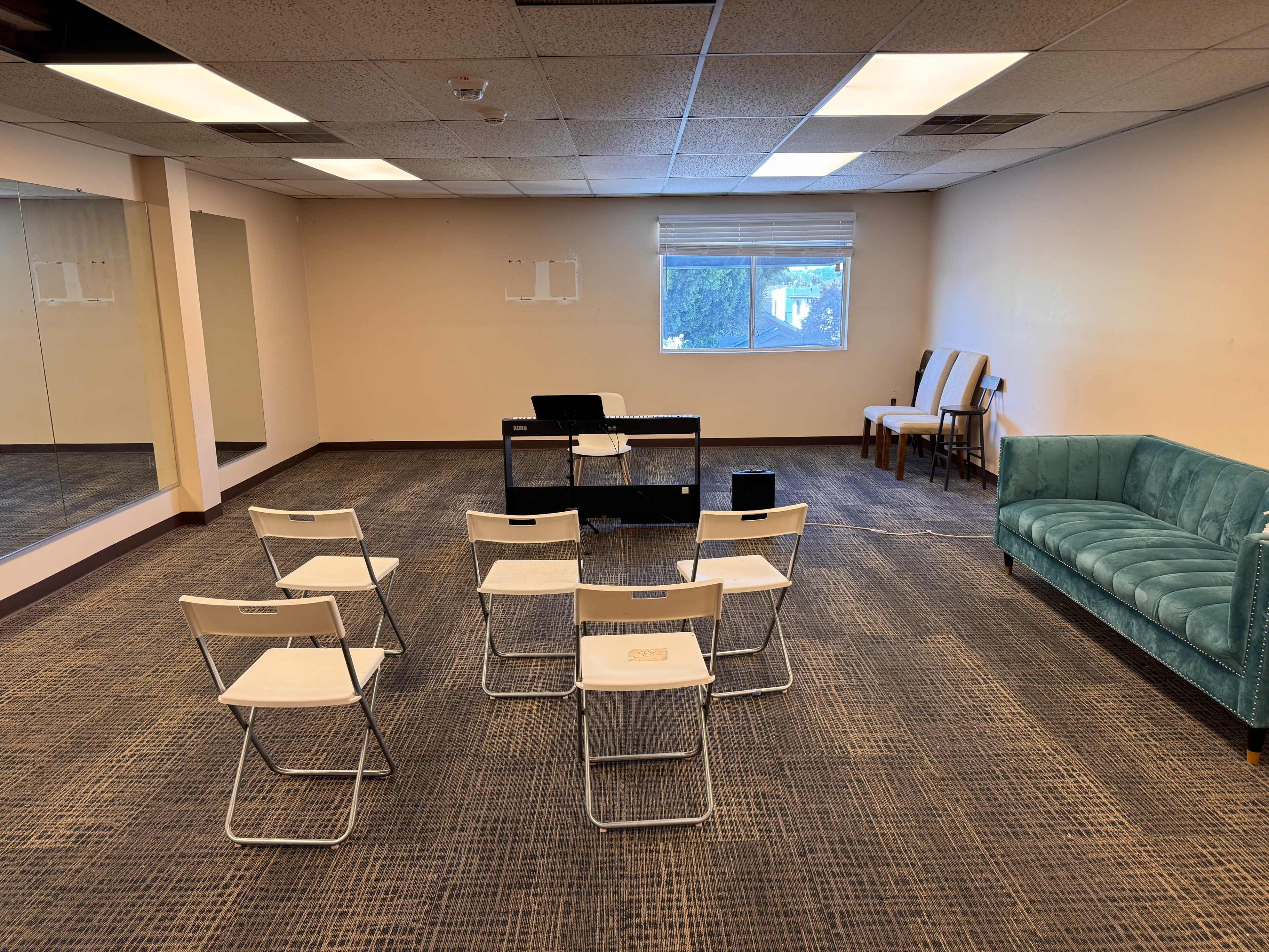 The image shows a sparse room with a few folding chairs arranged in a circle around a small table in front of a window, with a couch and additional chairs against one wall.