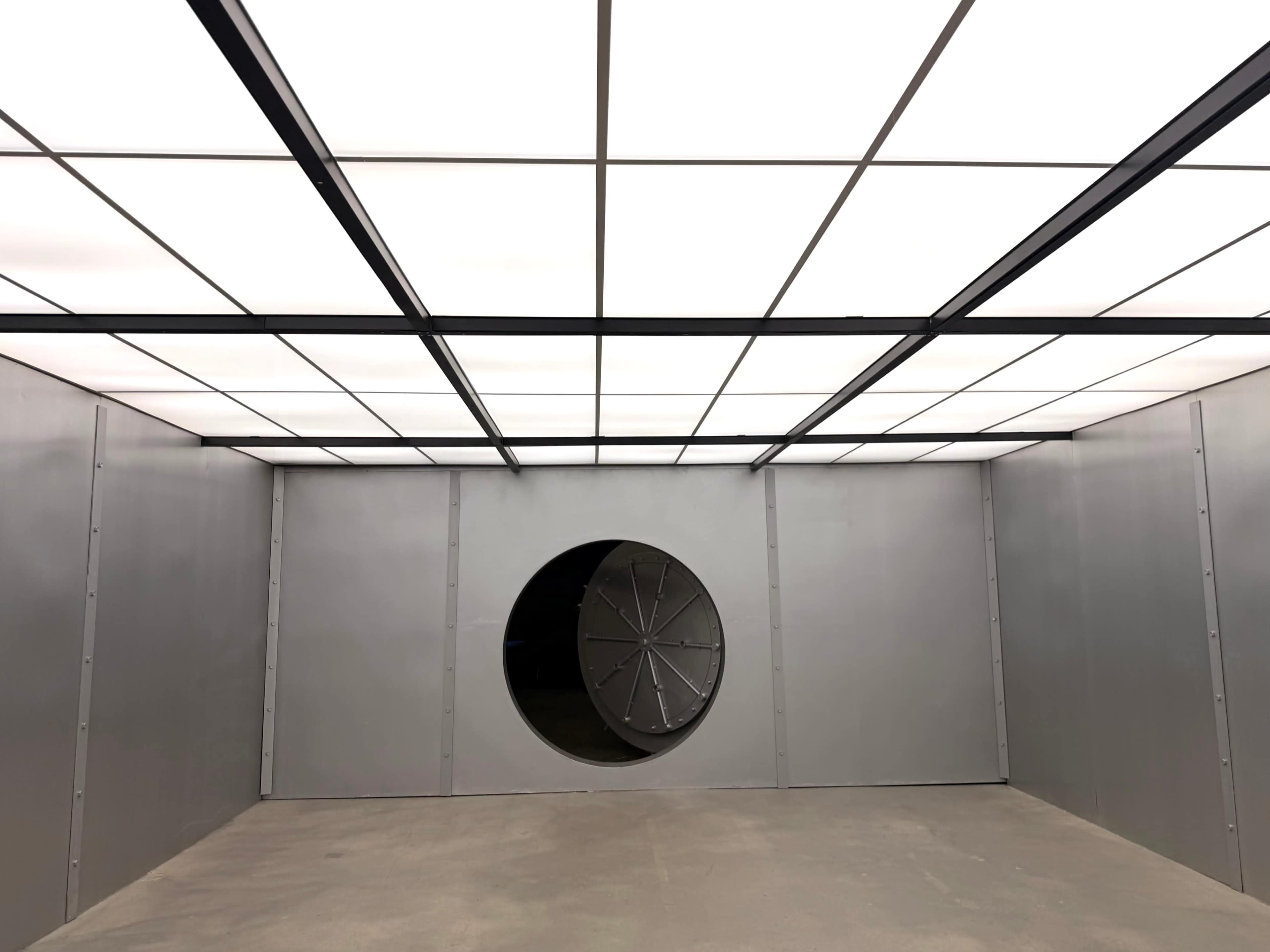 The image shows a large, empty room with gray walls and a circular vent on one side, illuminated by a grid of overhead lights.