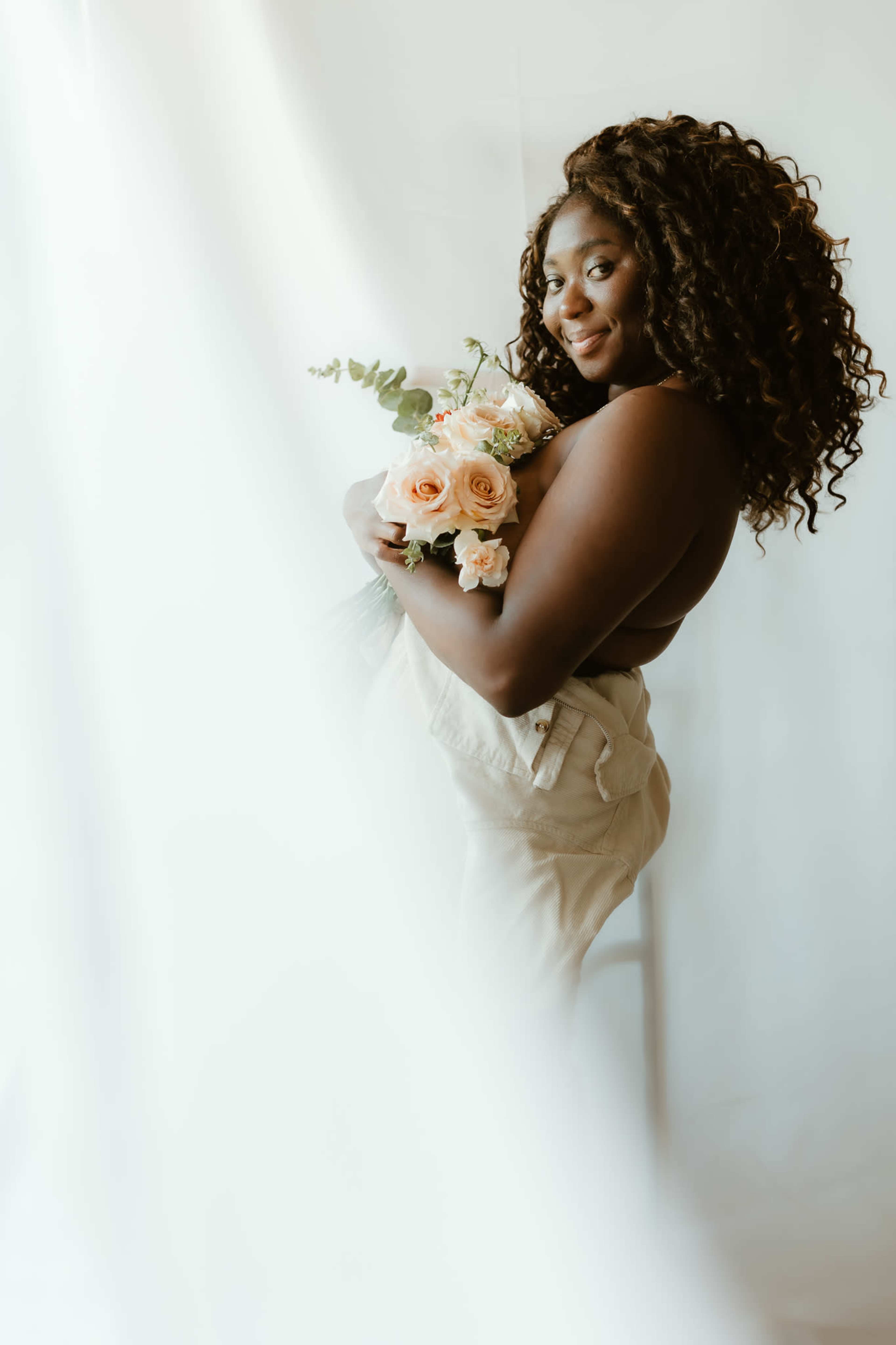 A woman with curly hair holds a bouquet of roses while standing against a light-colored backdrop.