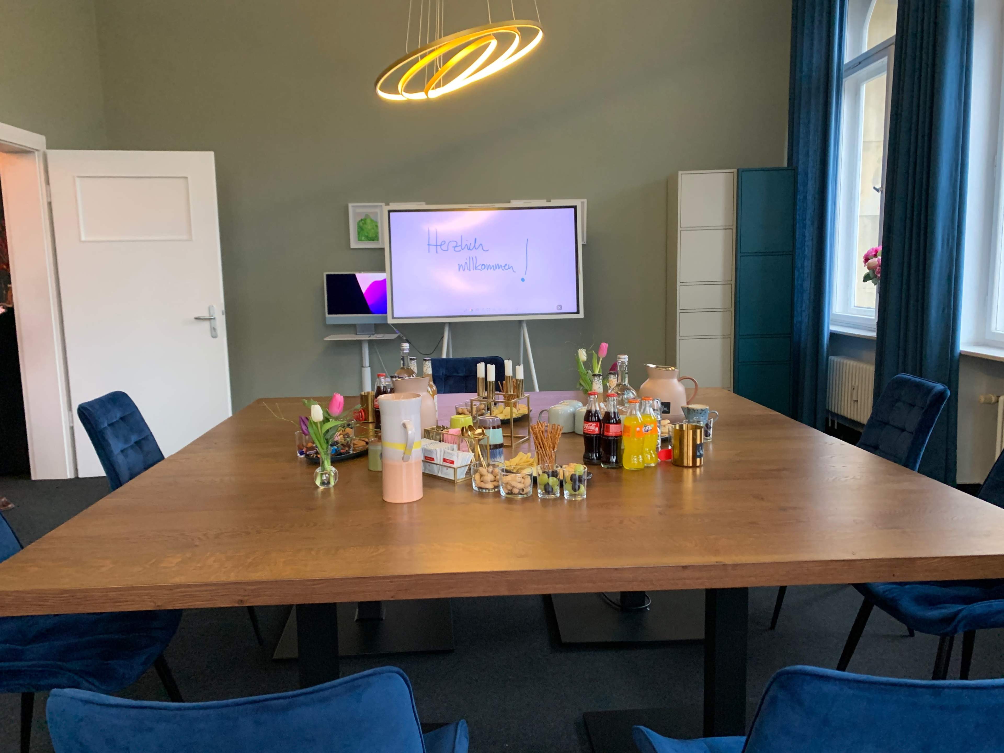 A conference room table is set up with various snacks and beverages, surrounded by blue chairs, and a presentation screen displays "Herzlich willkommen."