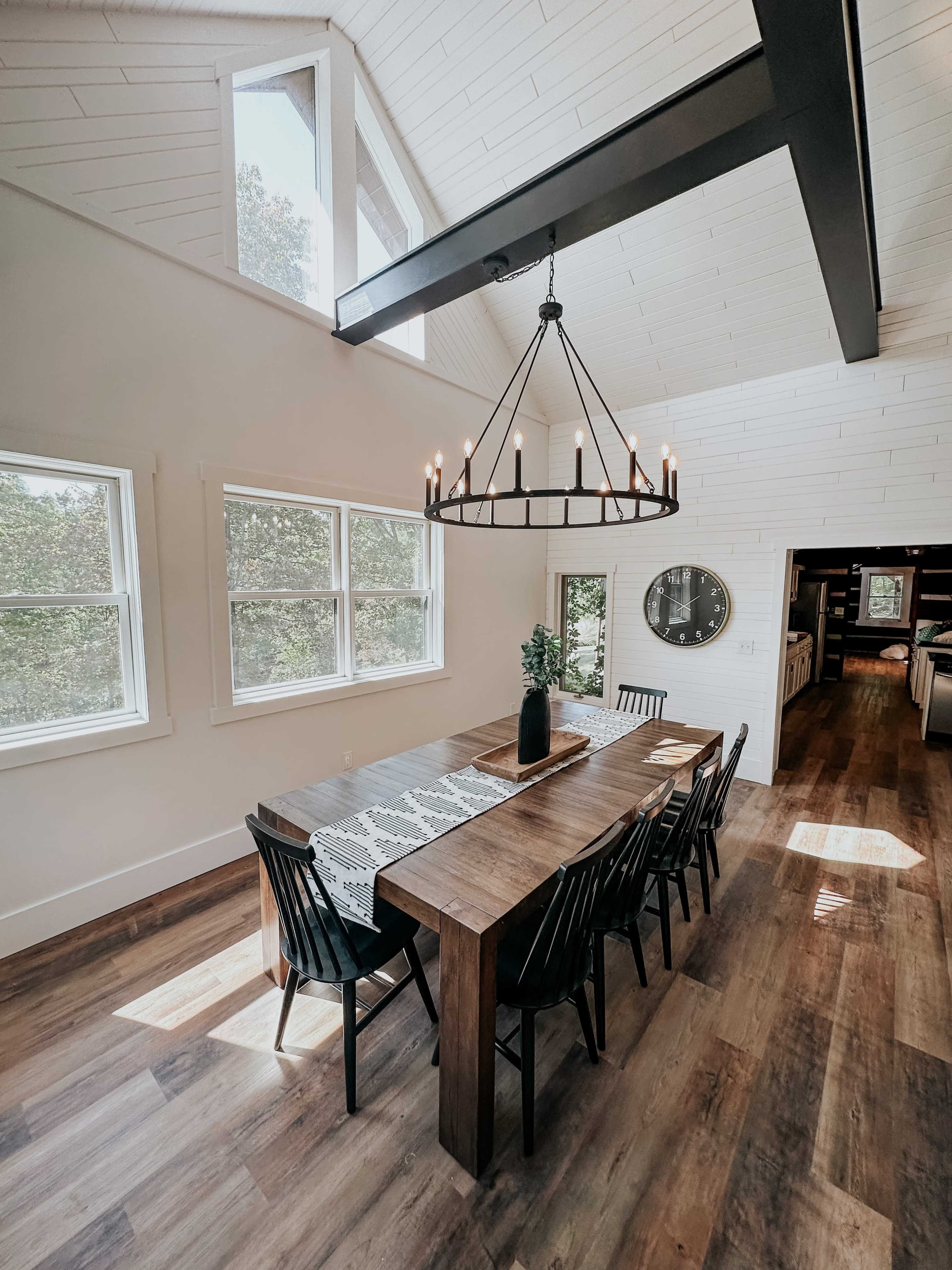 A dining area with a wooden table surrounded by black chairs, a large chandelier overhead, and bright windows letting in natural light.