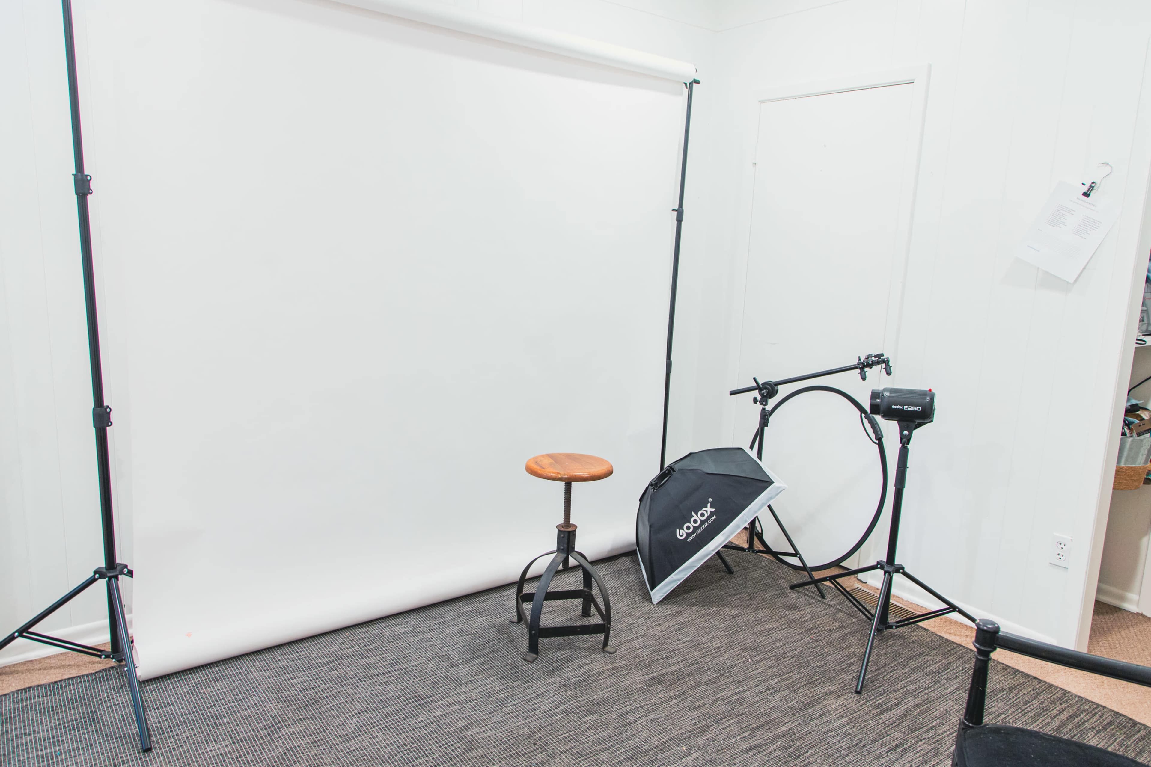 A photography studio setup featuring a plain white backdrop, a wooden stool, and lighting equipment in a corner space.