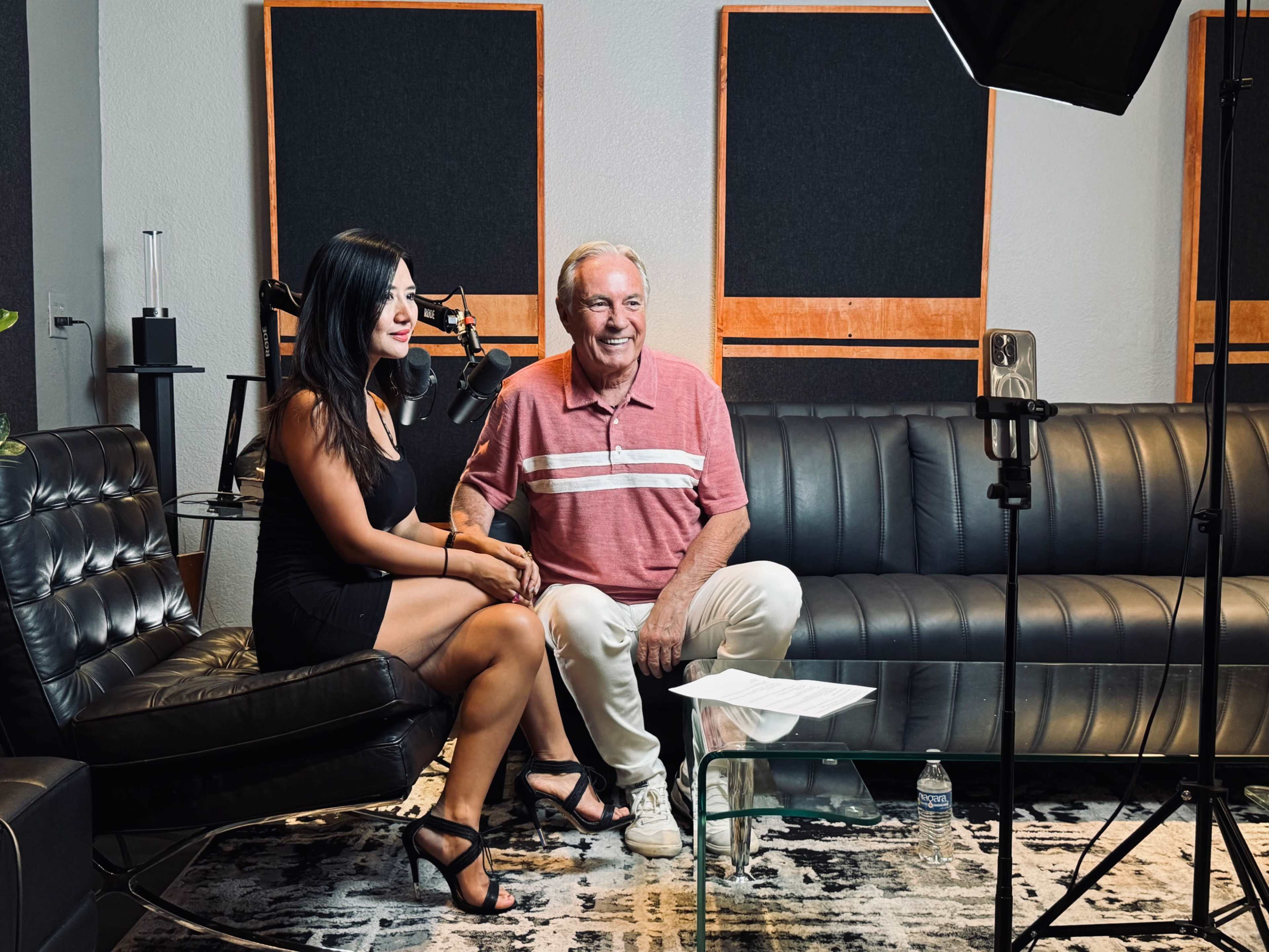 A man and a woman sit together on a leather sofa in a recording studio, with microphones positioned nearby.
