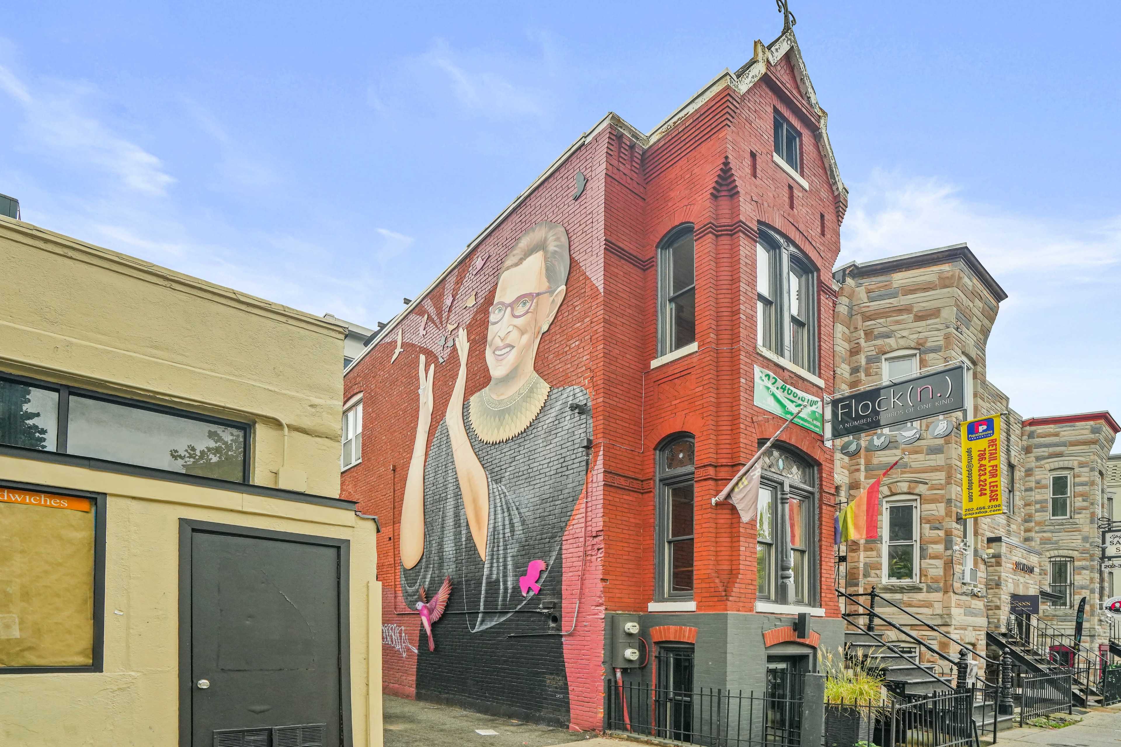 A large red brick building features a mural of a woman with glasses and dark hair, while a neighboring building displays a sign that reads "Flock(in)."