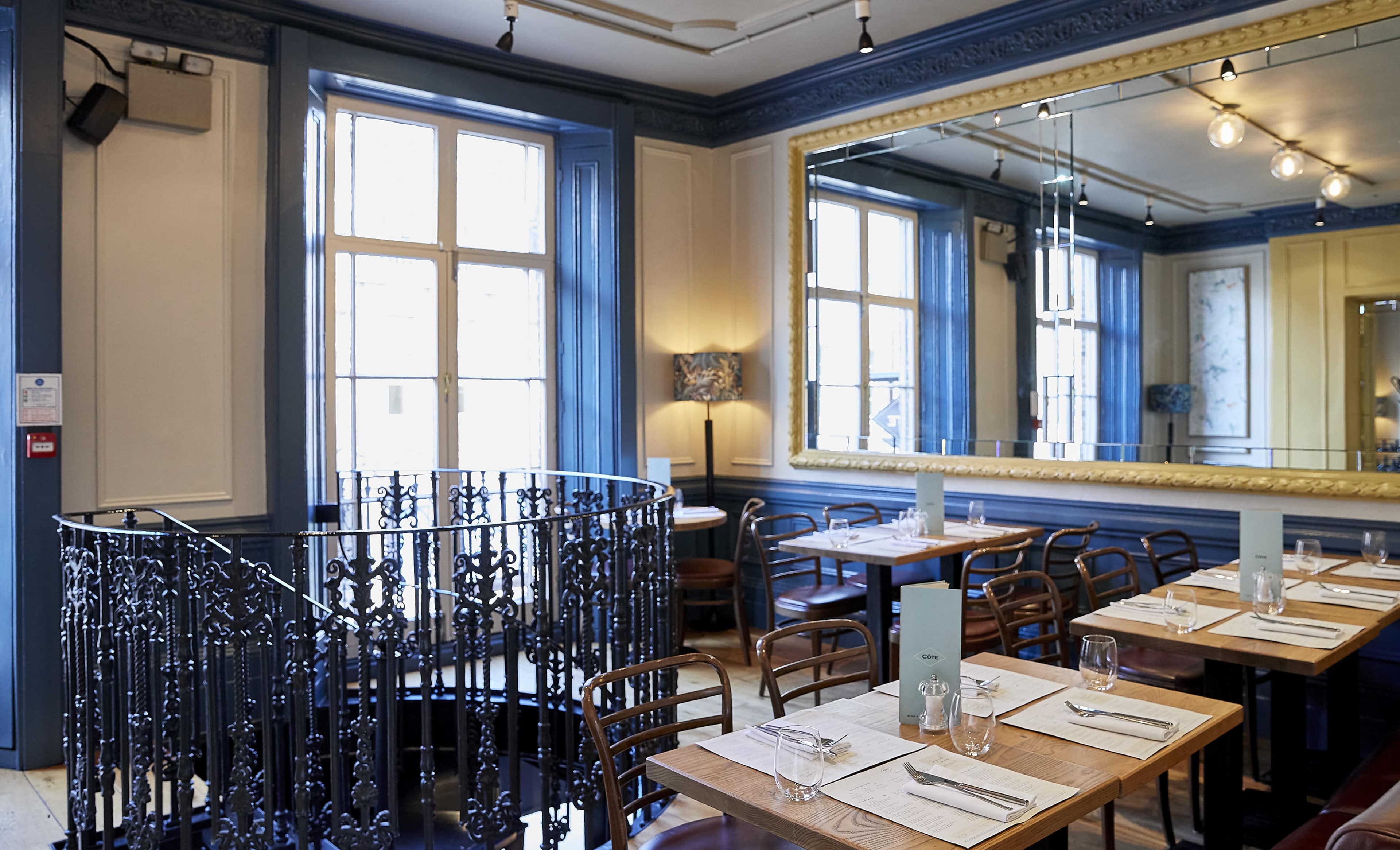 The image shows a restaurant interior with blue walls, wooden tables set with utensils, and a circular staircase leading to a lower level.