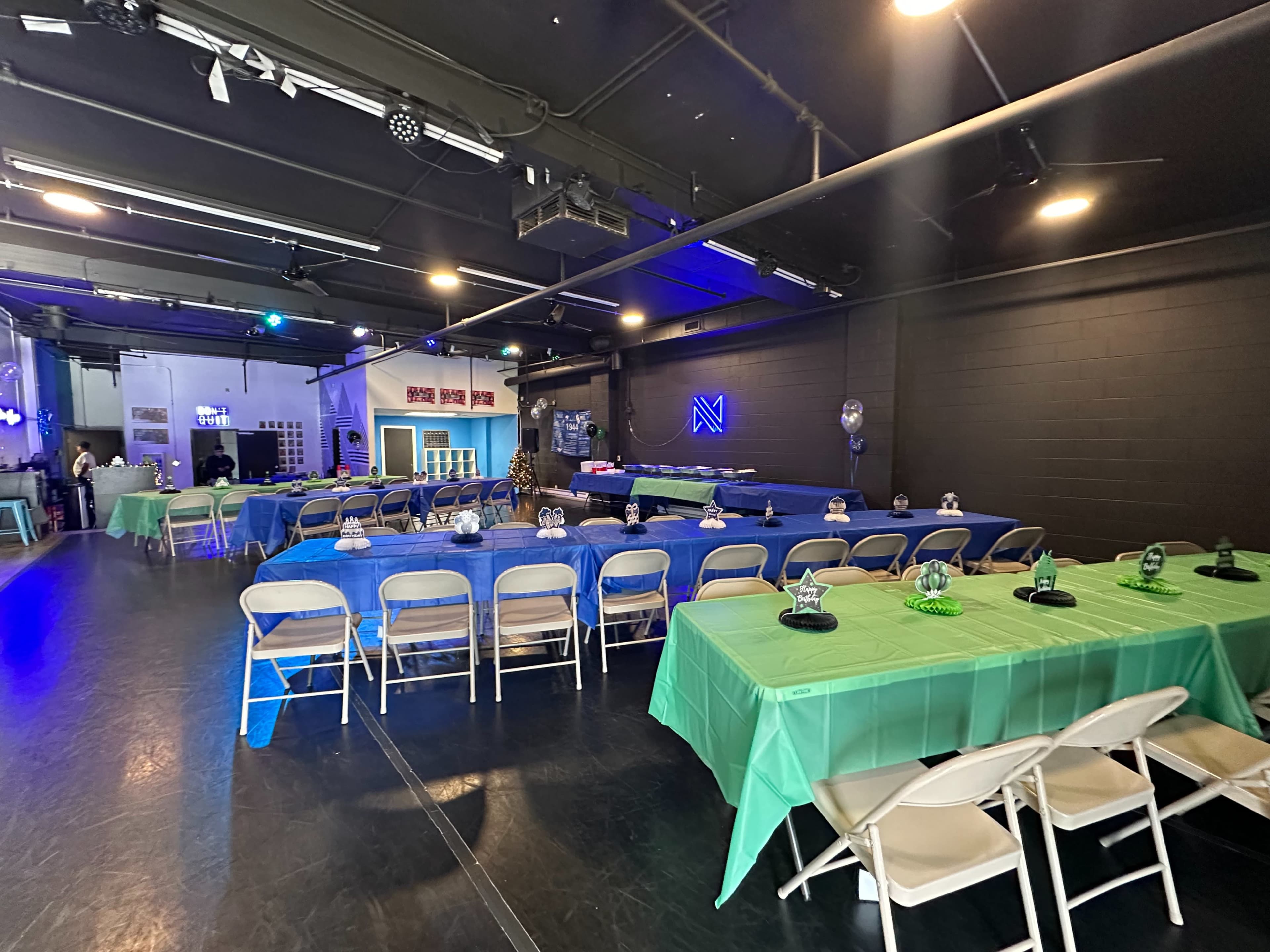 The image shows a spacious event room filled with long tables covered in blue and green table cloths, surrounded by folding chairs.