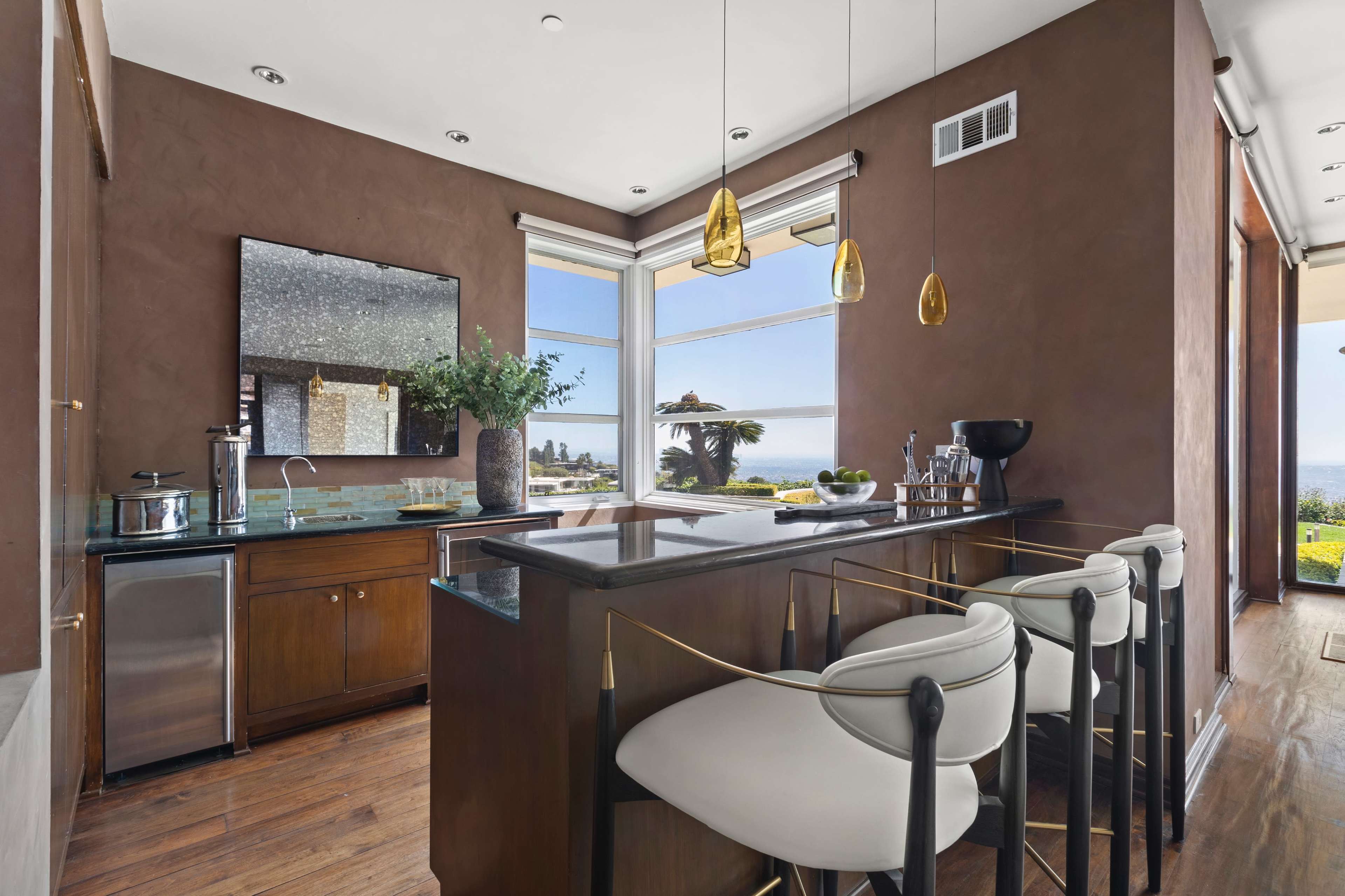 The image shows a modern kitchen bar area with a marble countertop, pendant lights, and a view of an outdoor landscape through large windows.