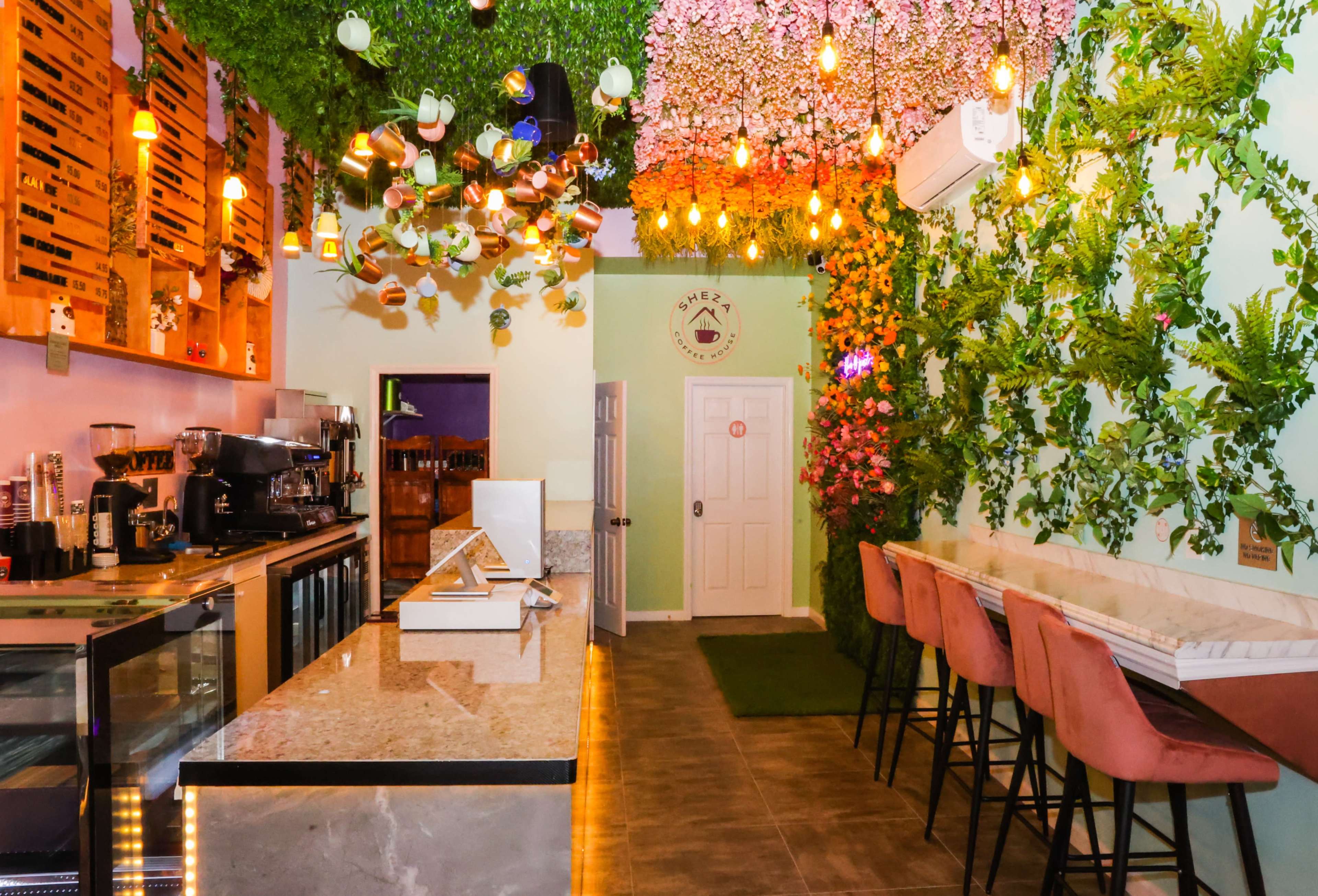 A modern café features a counter with a marble top, decorated walls covered in greenery and flowers, and colorful hanging light fixtures.