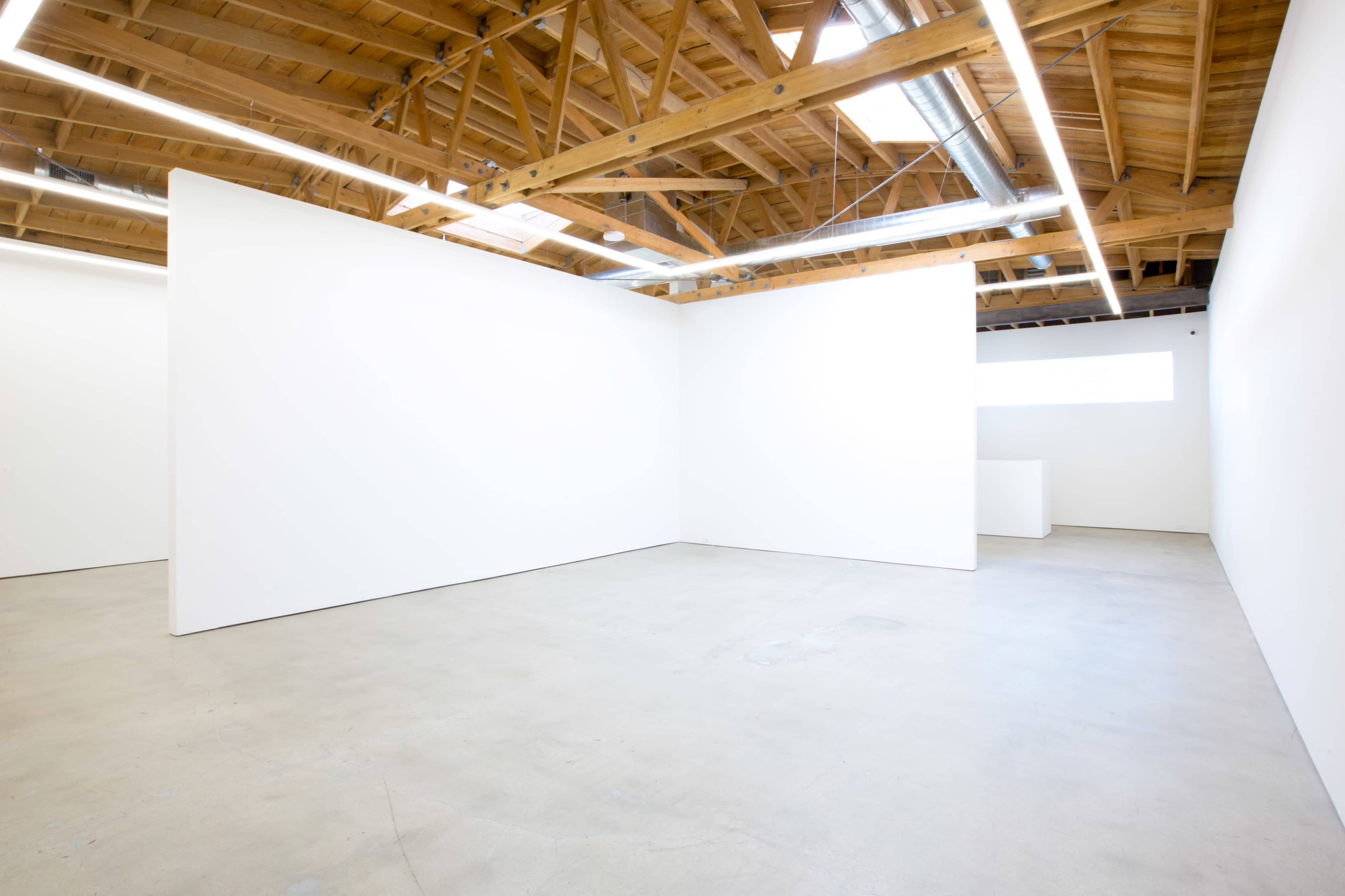The image shows a spacious, minimalist art gallery with white walls and a concrete floor, featuring wooden beams and lighting in the ceiling.