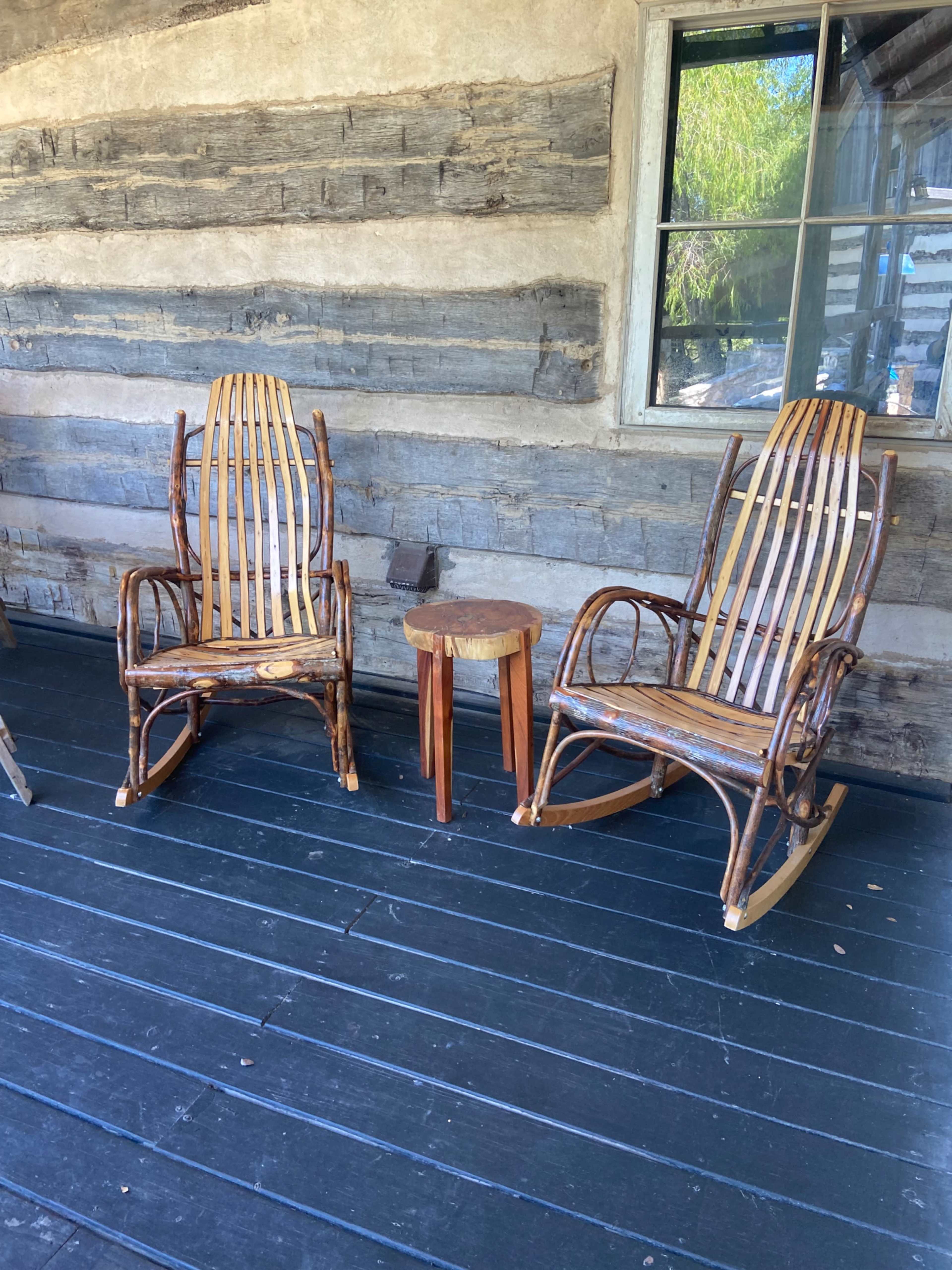 The image shows two wooden rocking chairs flanking a small wooden table on a dark wooden porch with a log cabin wall in the background.