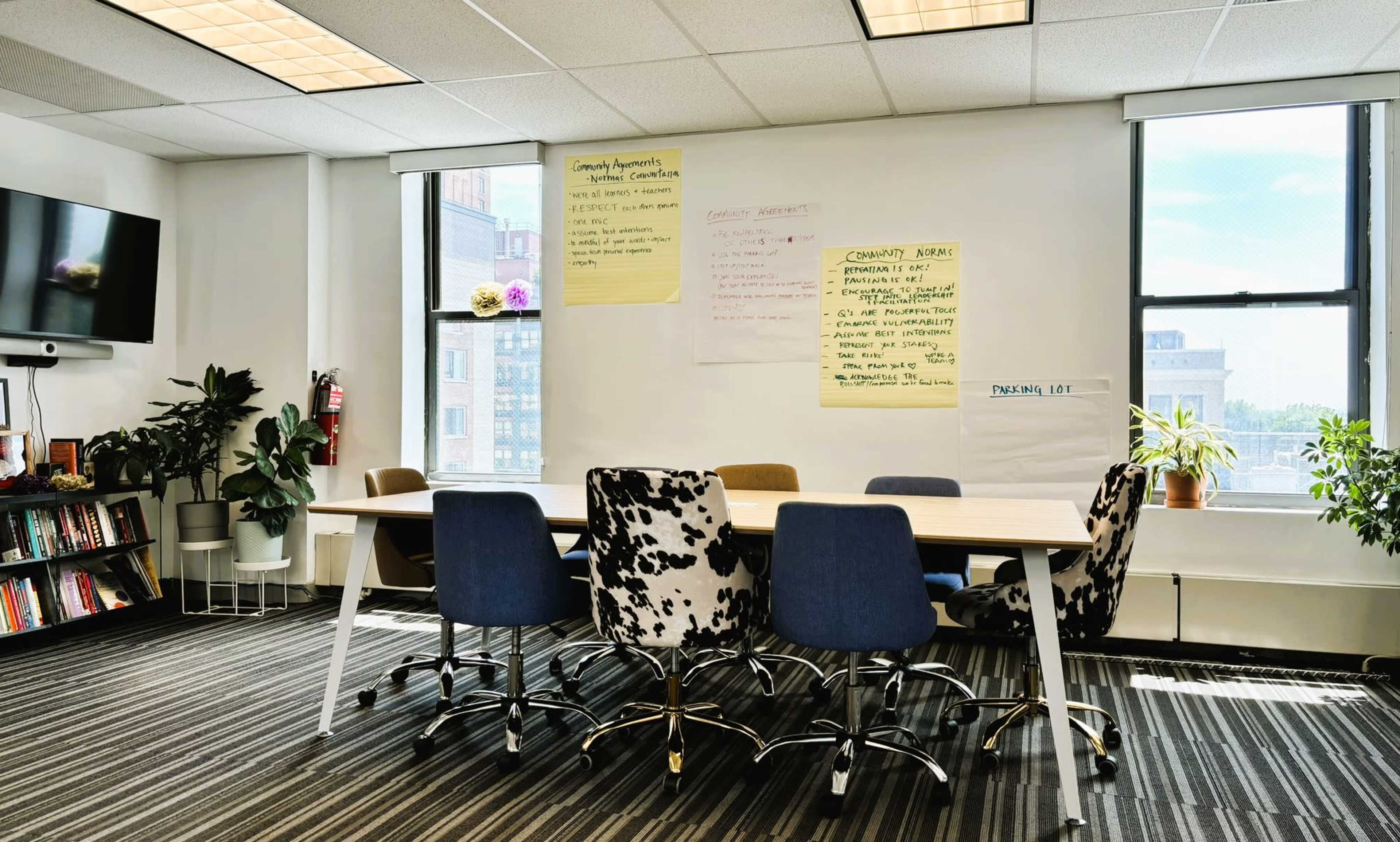 A meeting room features a long table surrounded by six chairs, with large windows and flip chart paper on the walls.