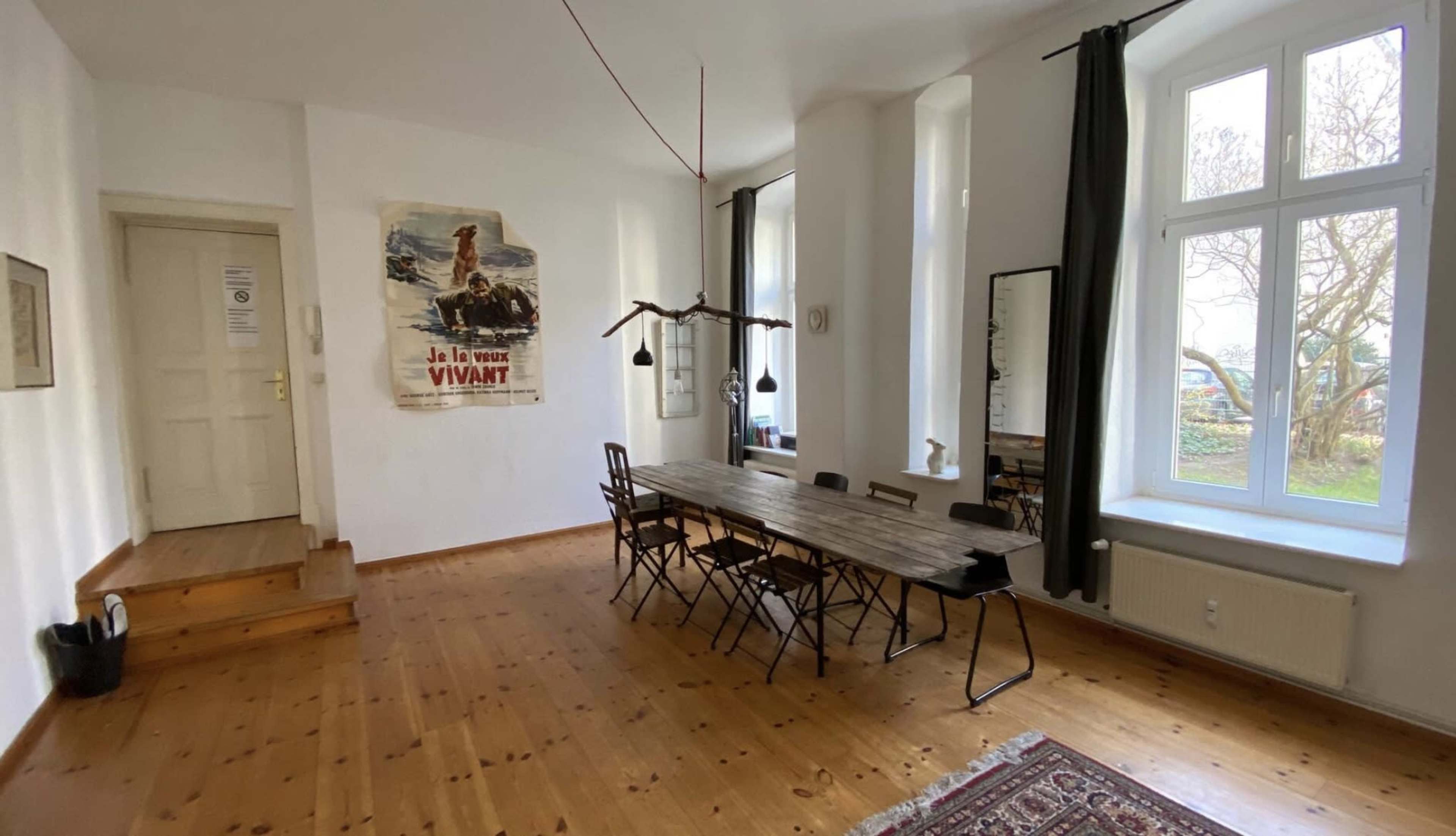 A spacious room featuring a long wooden dining table surrounded by chairs, with a large window allowing natural light and a poster hanging on the wall.