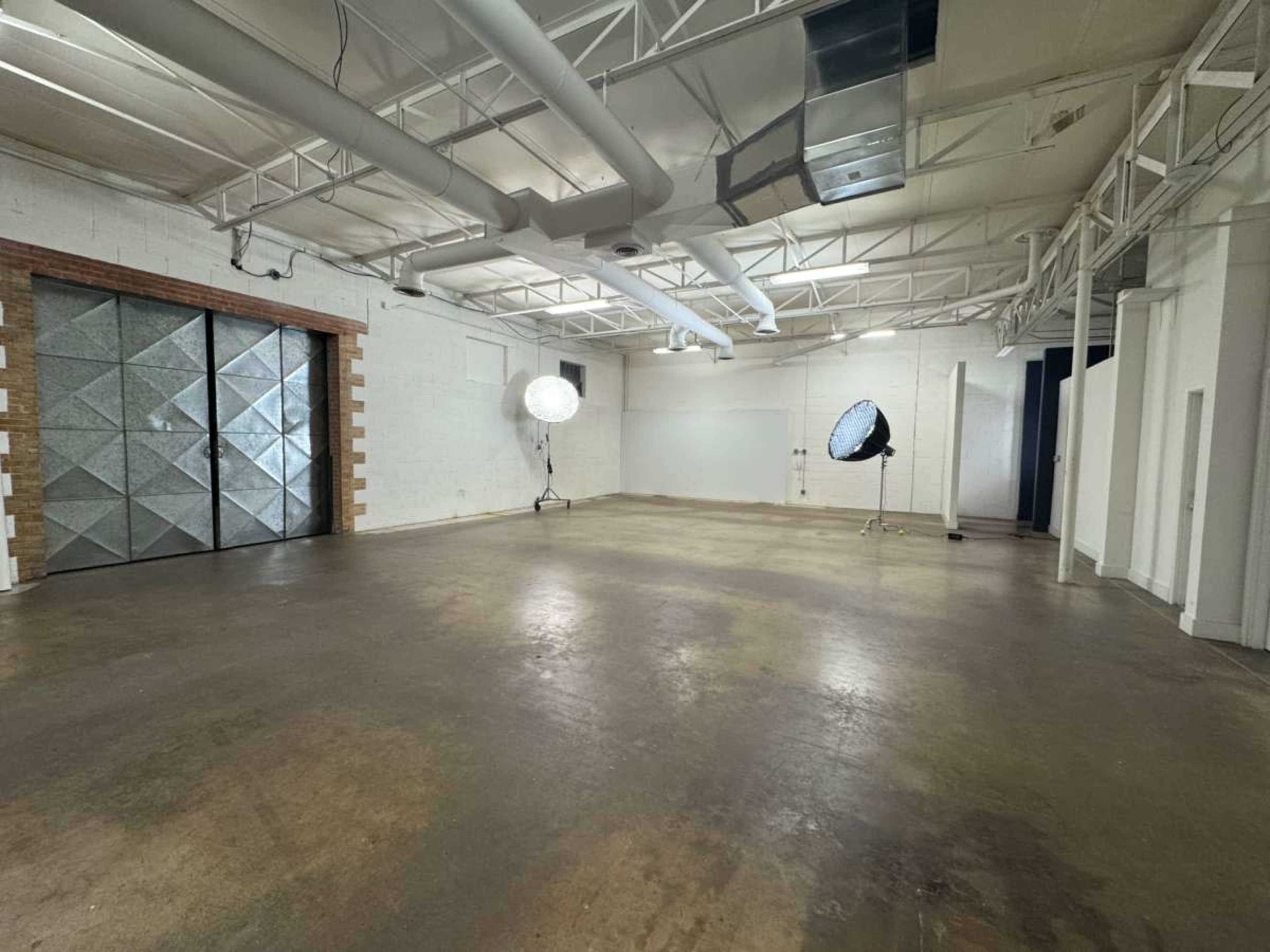 The image shows an empty industrial-style room with a concrete floor, illuminated by large studio lights, and featuring a textured metal door on one wall.
