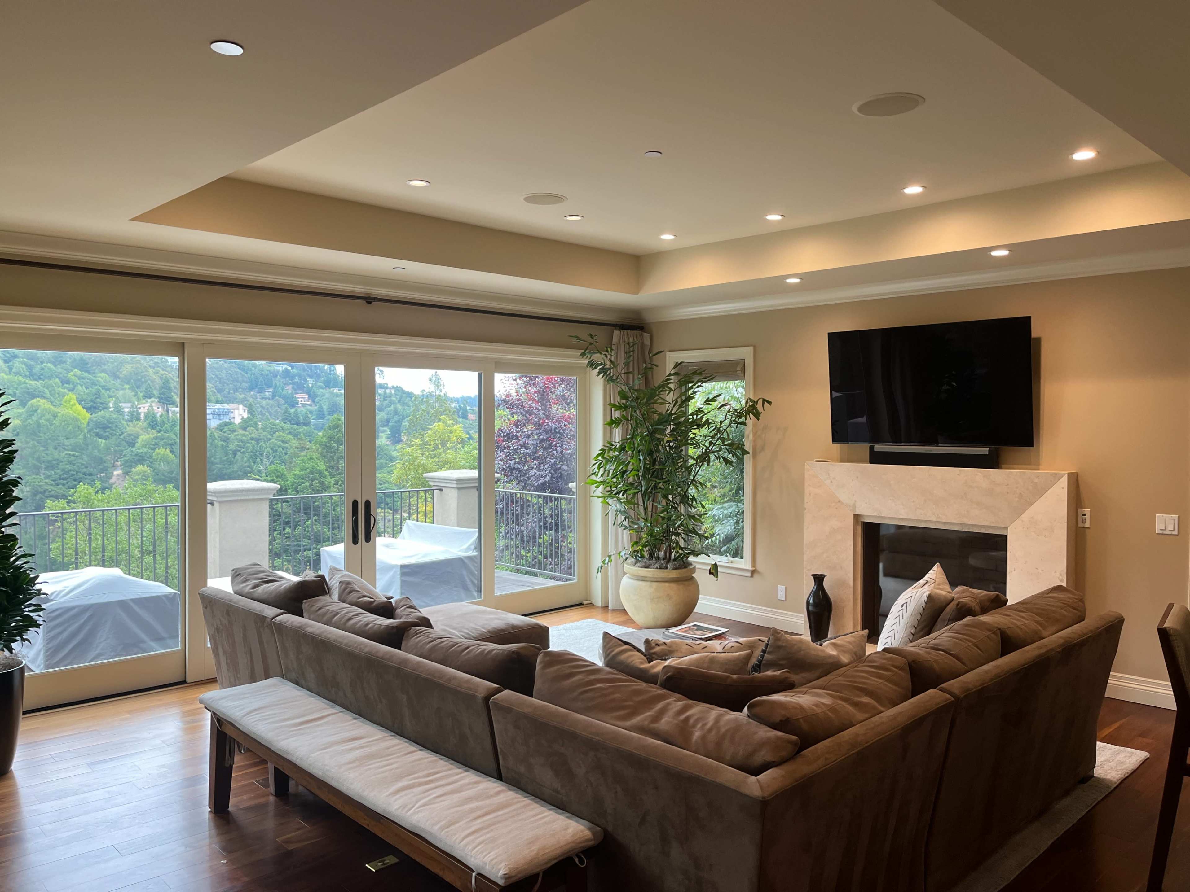 The image shows a spacious living room with a large sectional sofa, a TV mounted on the wall, and sliding glass doors leading to a balcony with a scenic view.