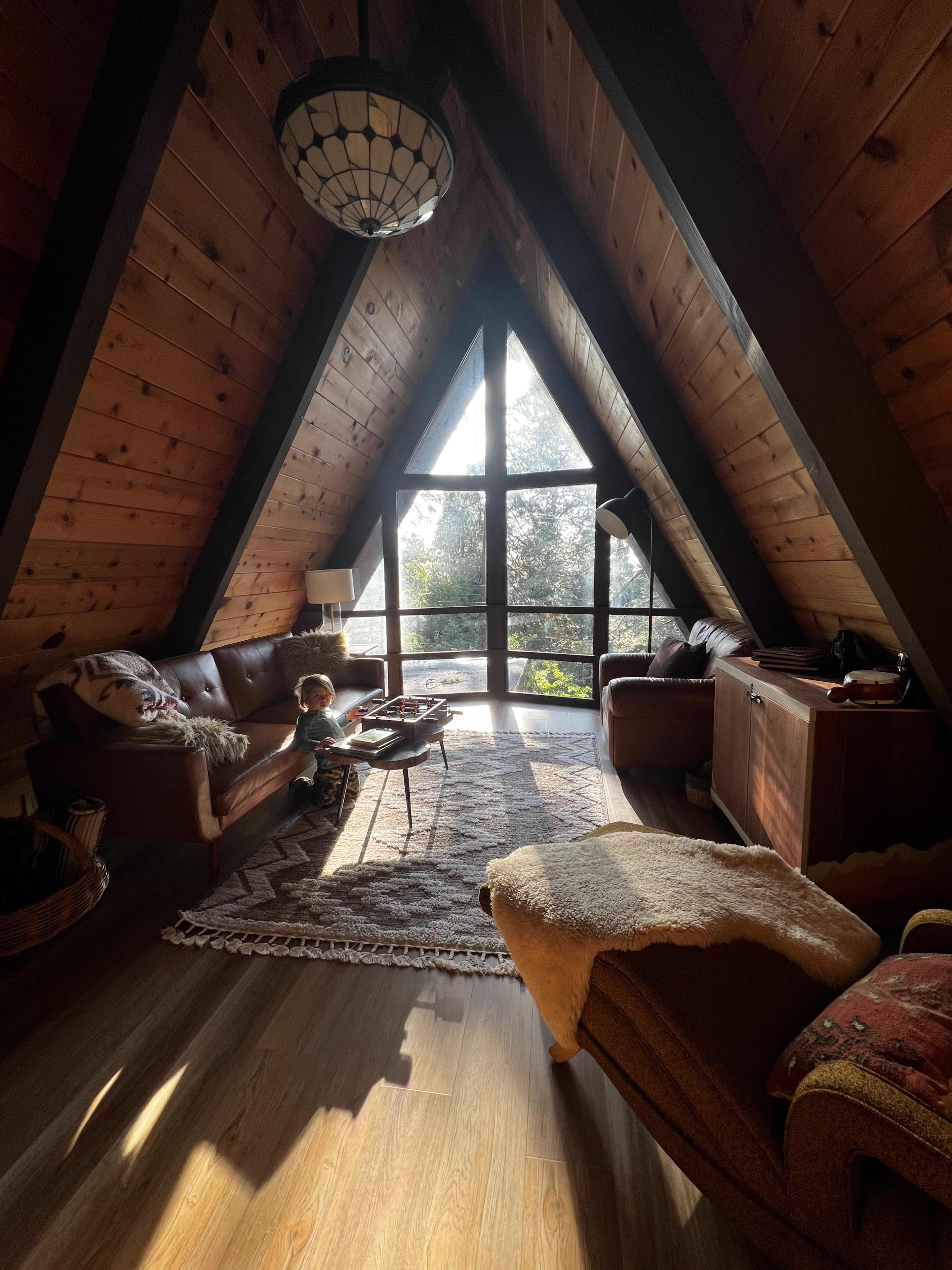 A child stands by a coffee table in a cozy, wooden A-frame living room with large windows letting in natural light.