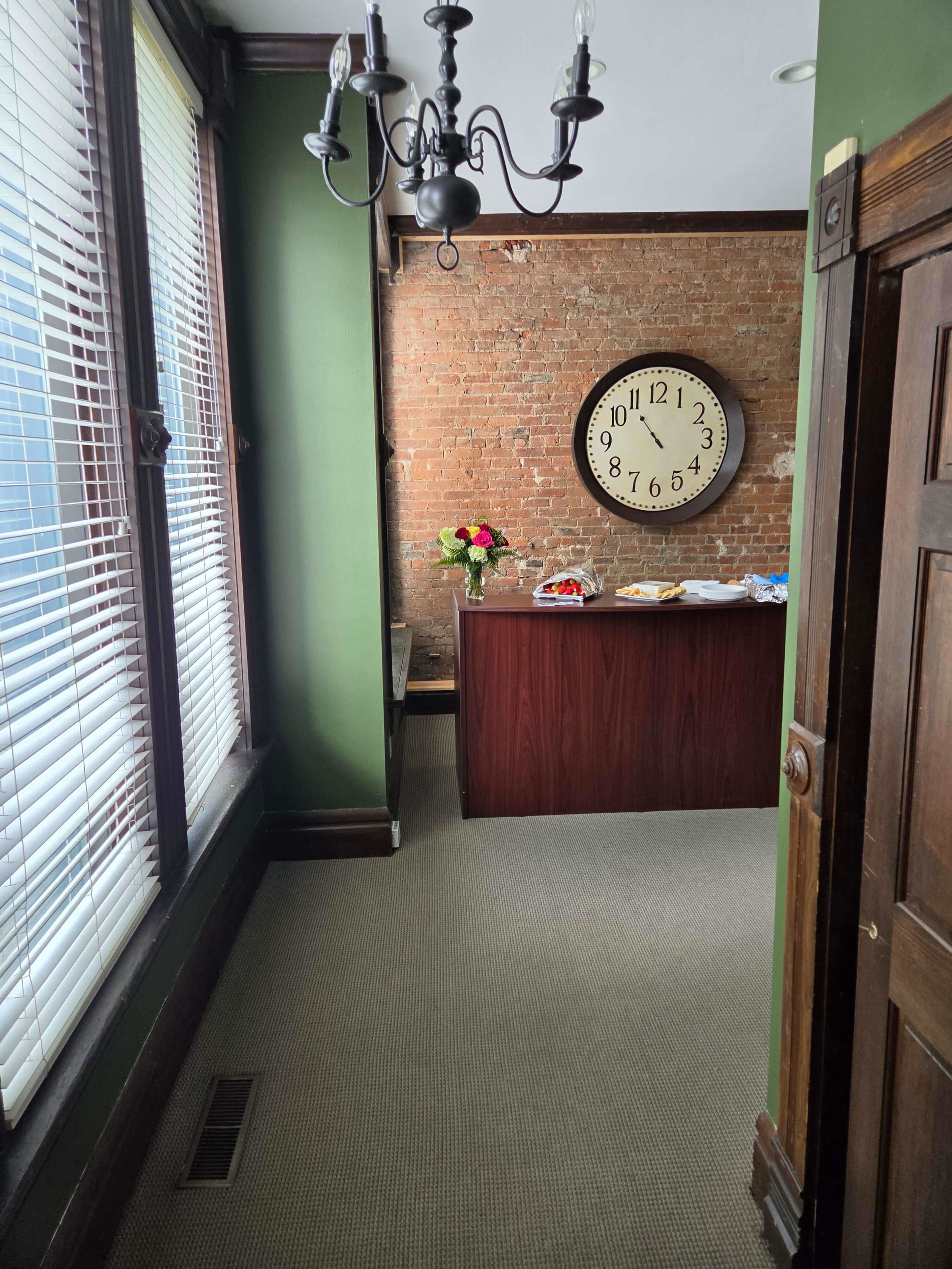 The image shows a corridor leading to a room with a brick wall, a large clock, a wooden reception desk, and a bouquet of flowers on the desk.
