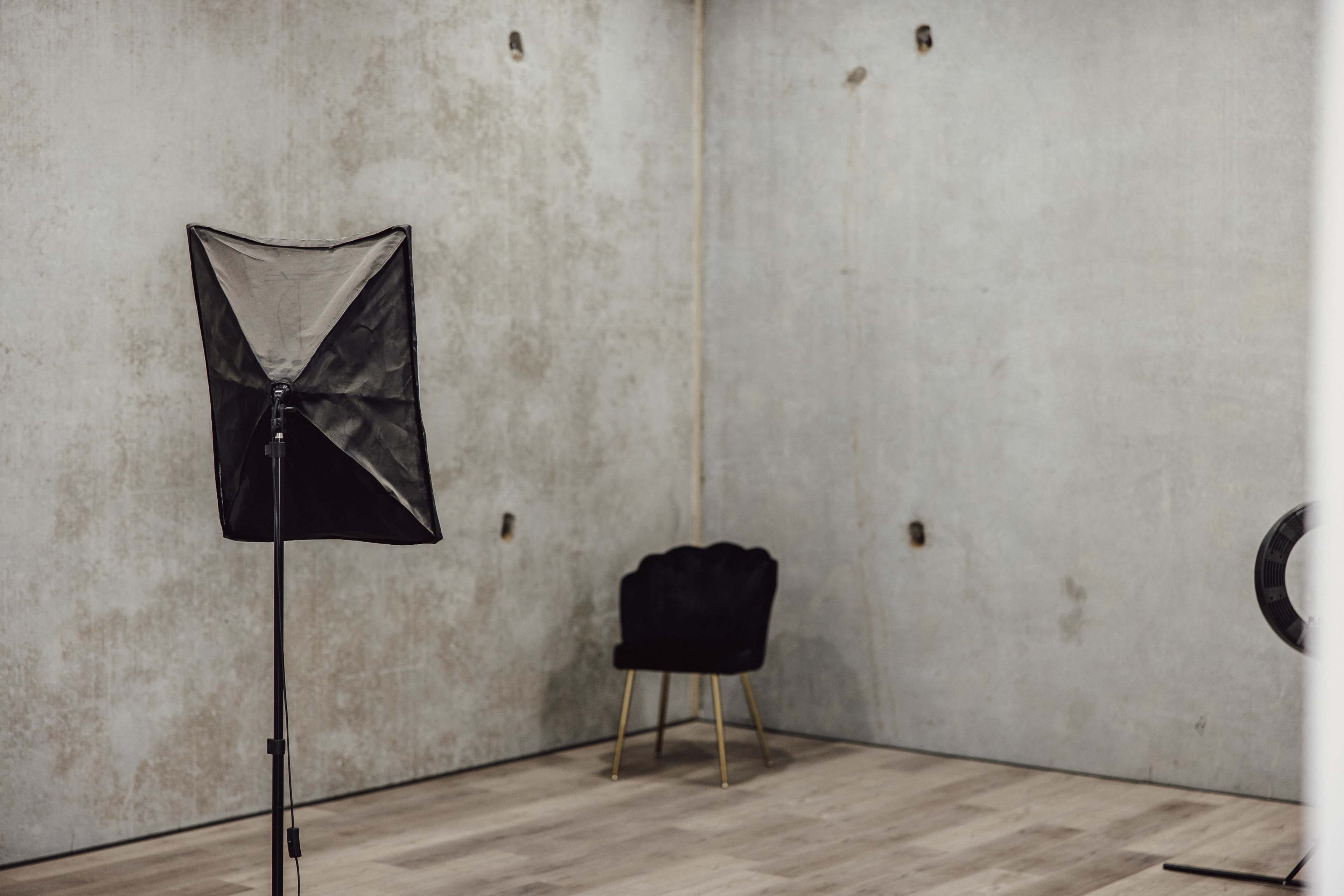 An empty room with a concrete wall, a black photography softbox on a stand, and a black chair with gold legs.