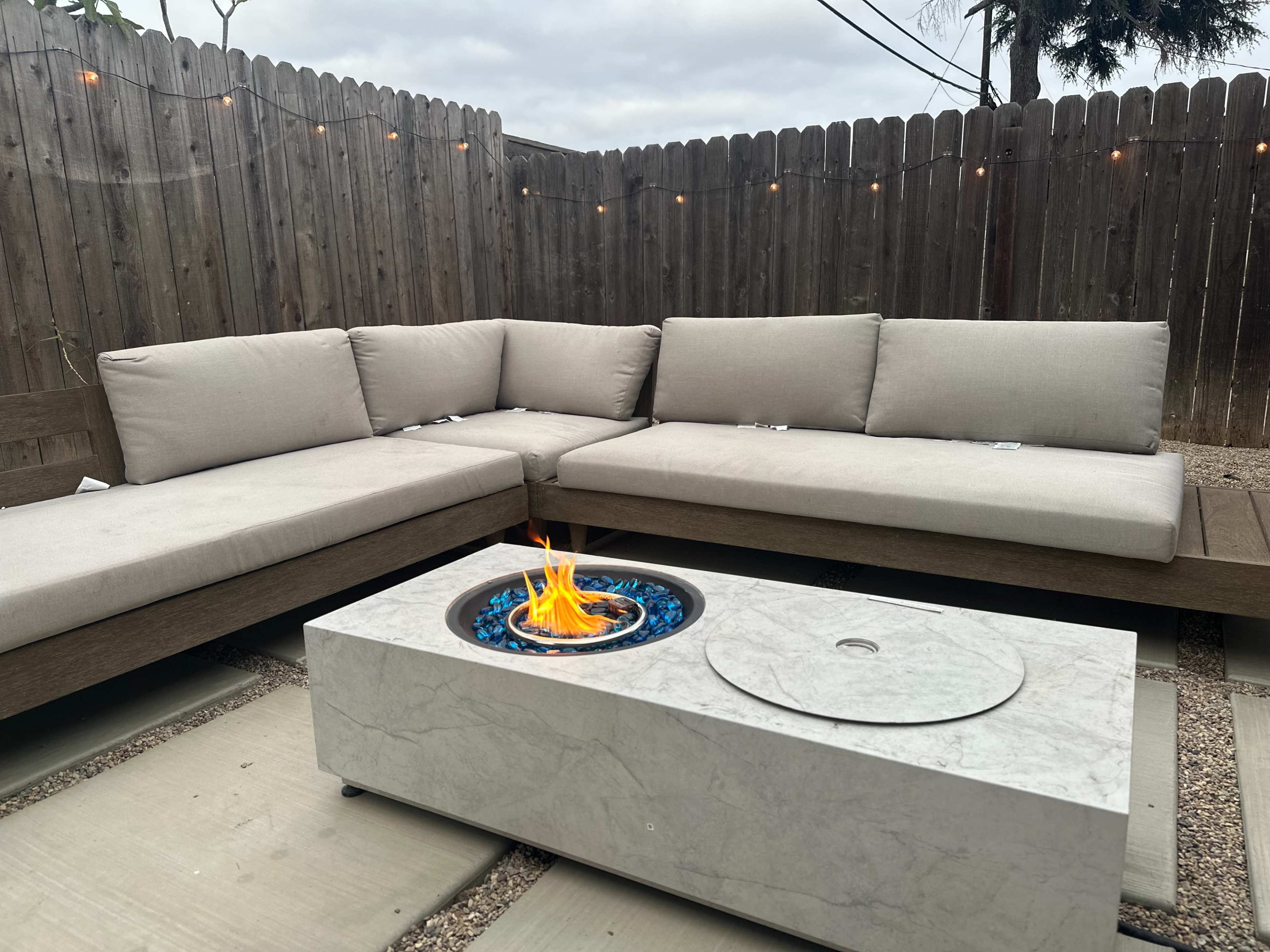 A modern outdoor seating area features a curved sectional couch with beige cushions and a stone fire pit with blue glass stones, surrounded by a wooden fence.