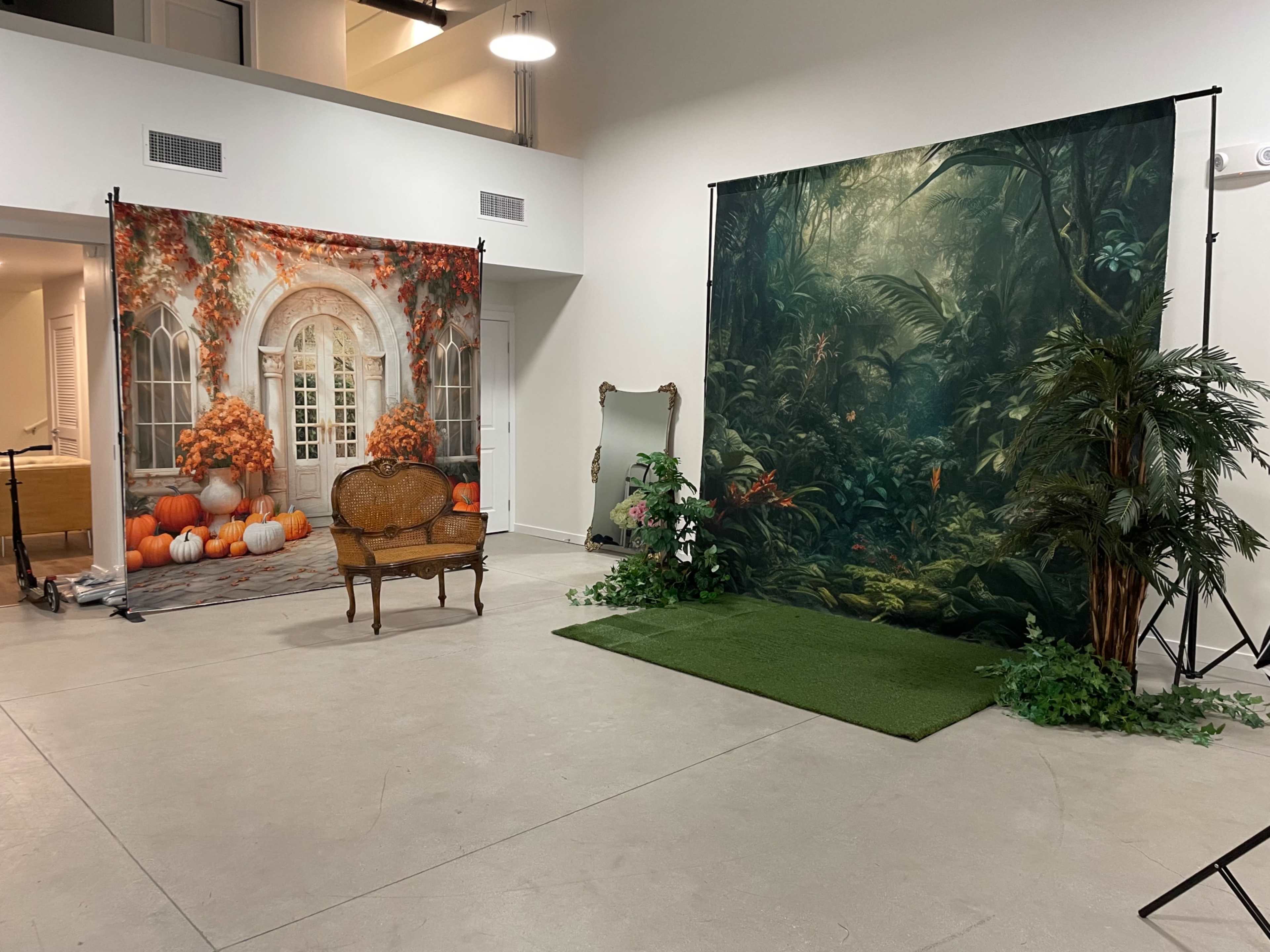 A spacious room with two distinct backdrop scenes: one with an autumn-themed setting featuring pumpkins and flowers, and the other depicting a lush jungle landscape, accompanied by a vintage chair and a decorative mirror.