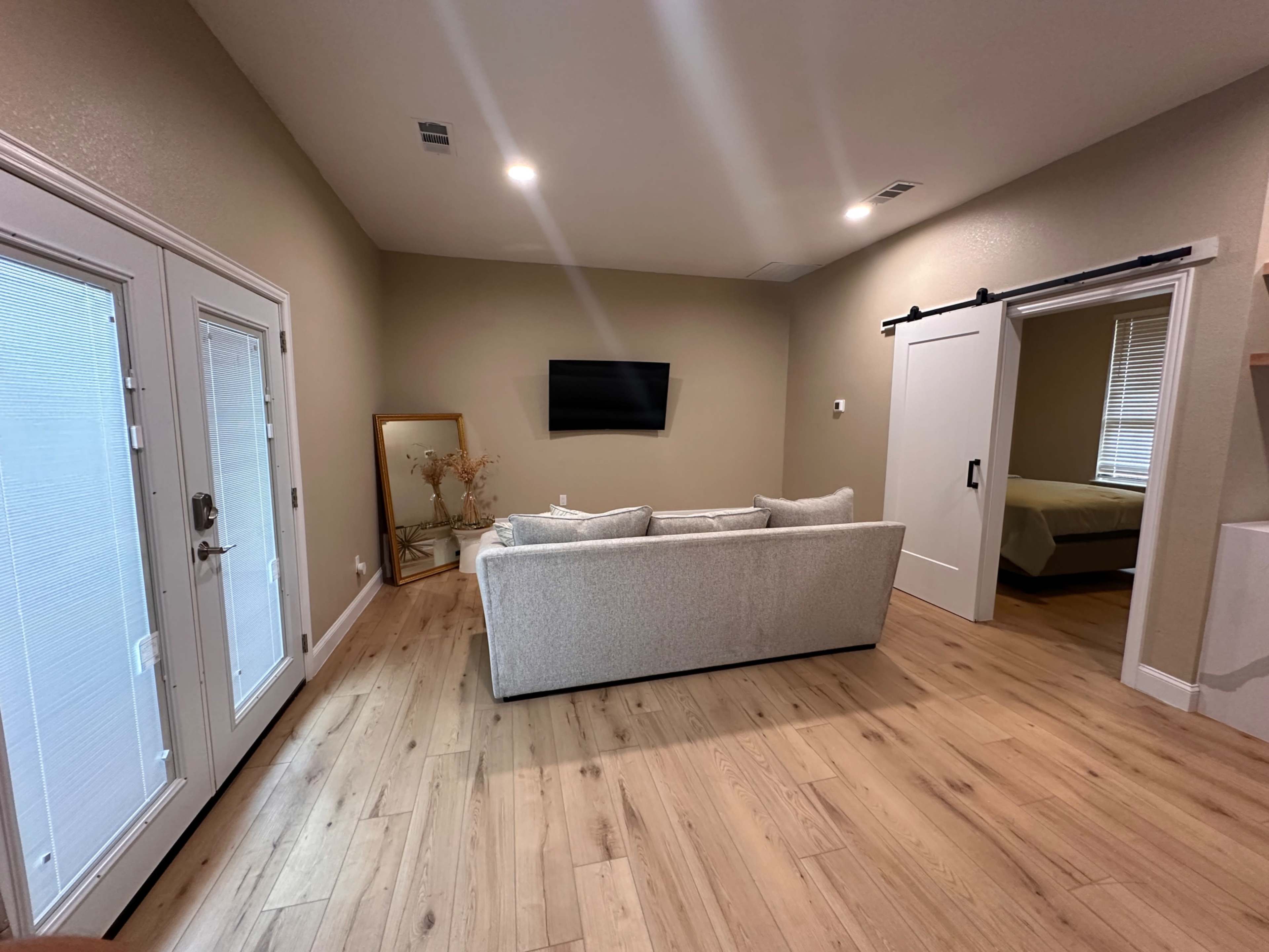 A living room featuring a light-colored sofa, a wall-mounted television, and large windows with natural light, complemented by wooden flooring.