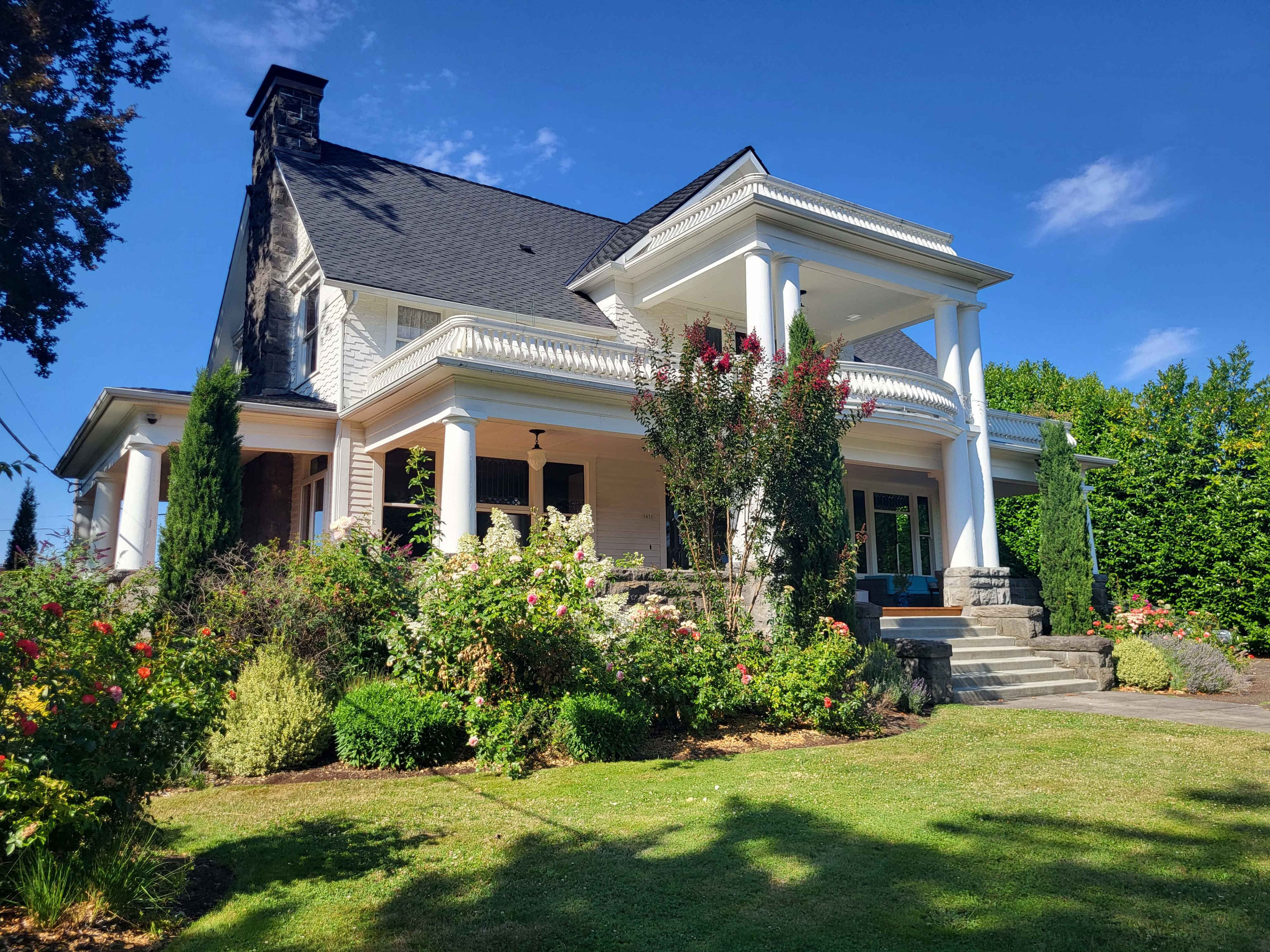 A large, two-story house with a front porch supported by white pillars, surrounded by landscaped gardens.