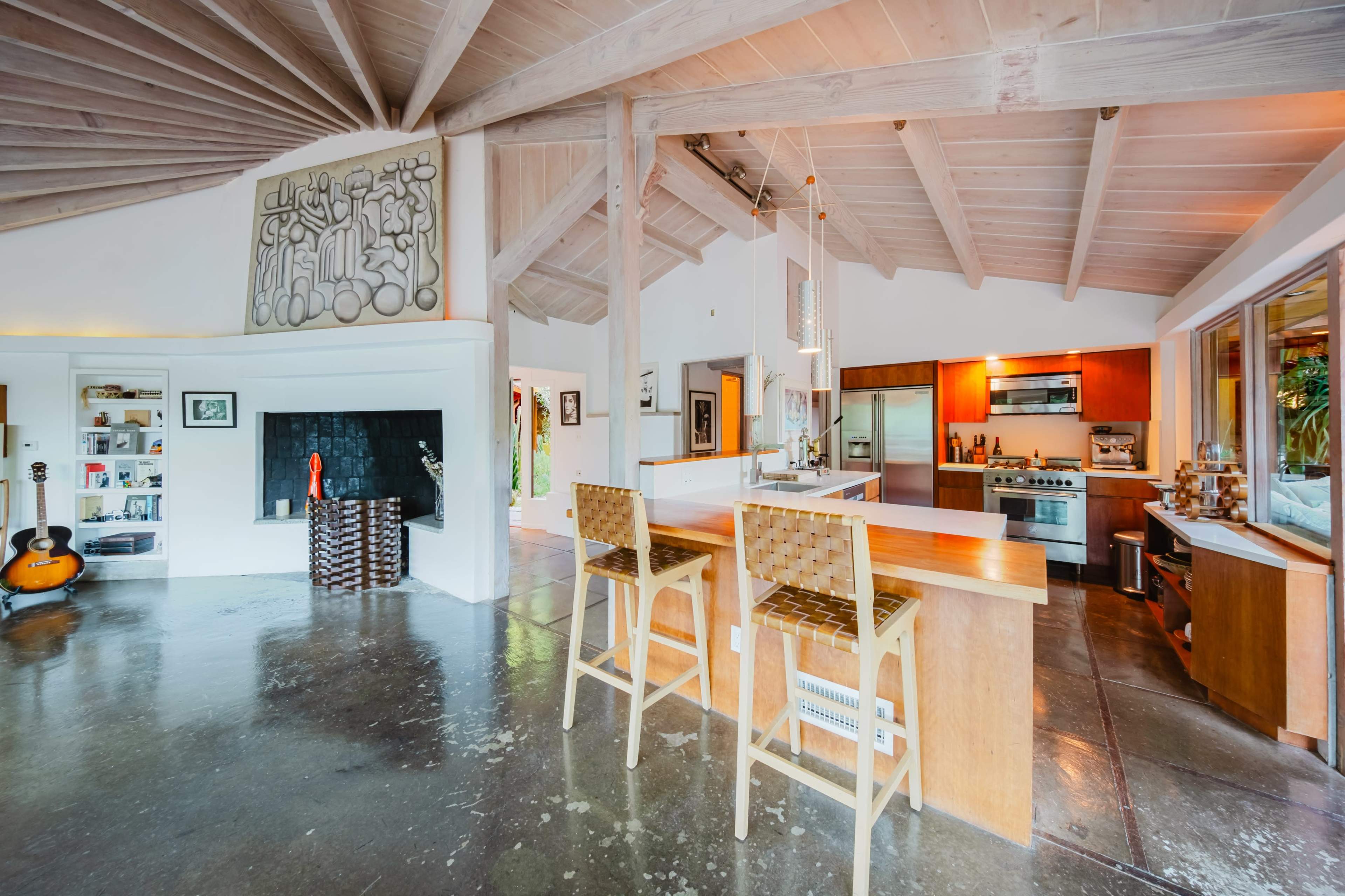 The image showcases a modern, open-concept kitchen and living space with a high, angled ceiling, highlighting wooden beams and a central island with two bar stools.