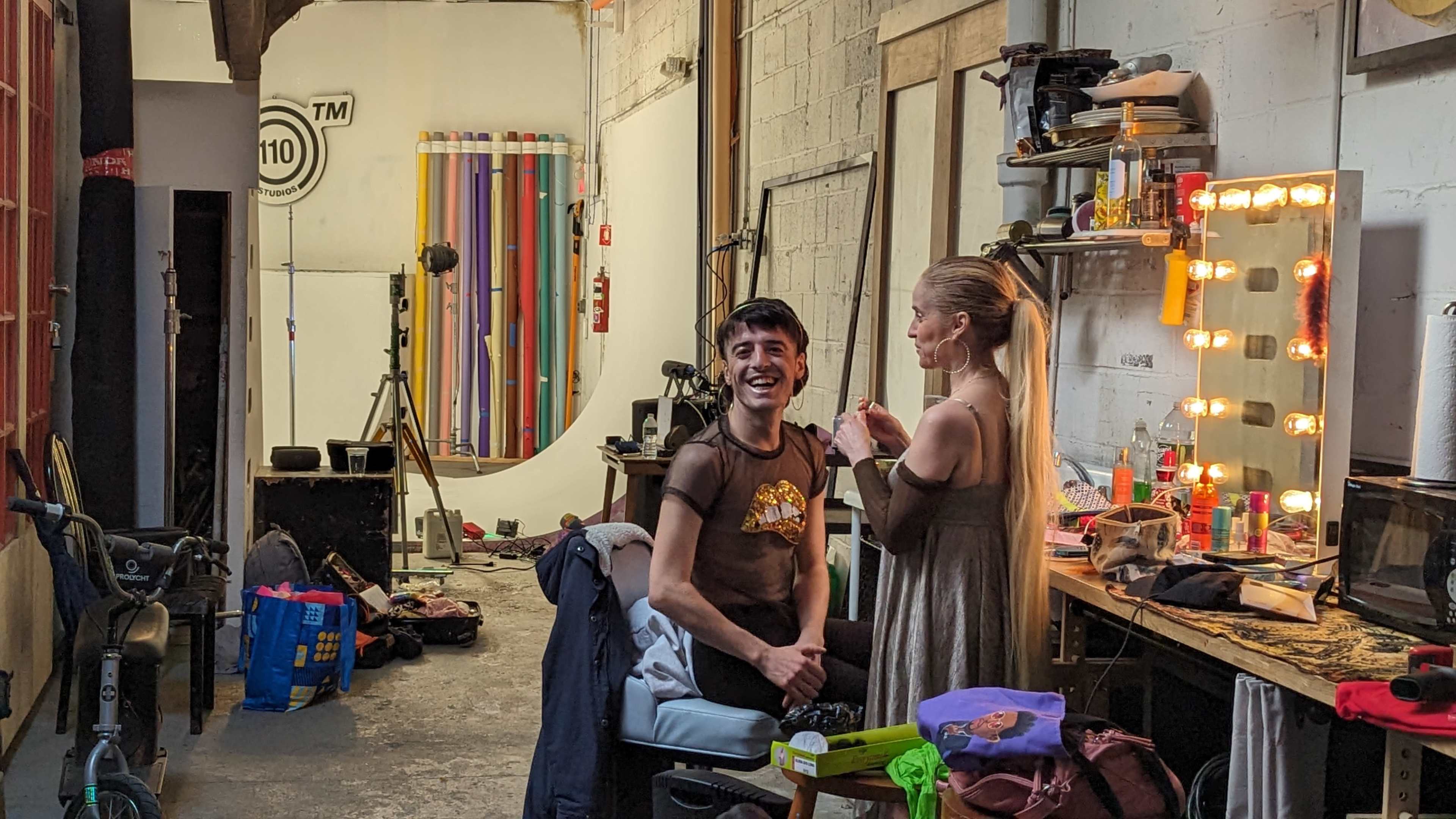 A person is seated in a studio while another person applies makeup, surrounded by various props and equipment.