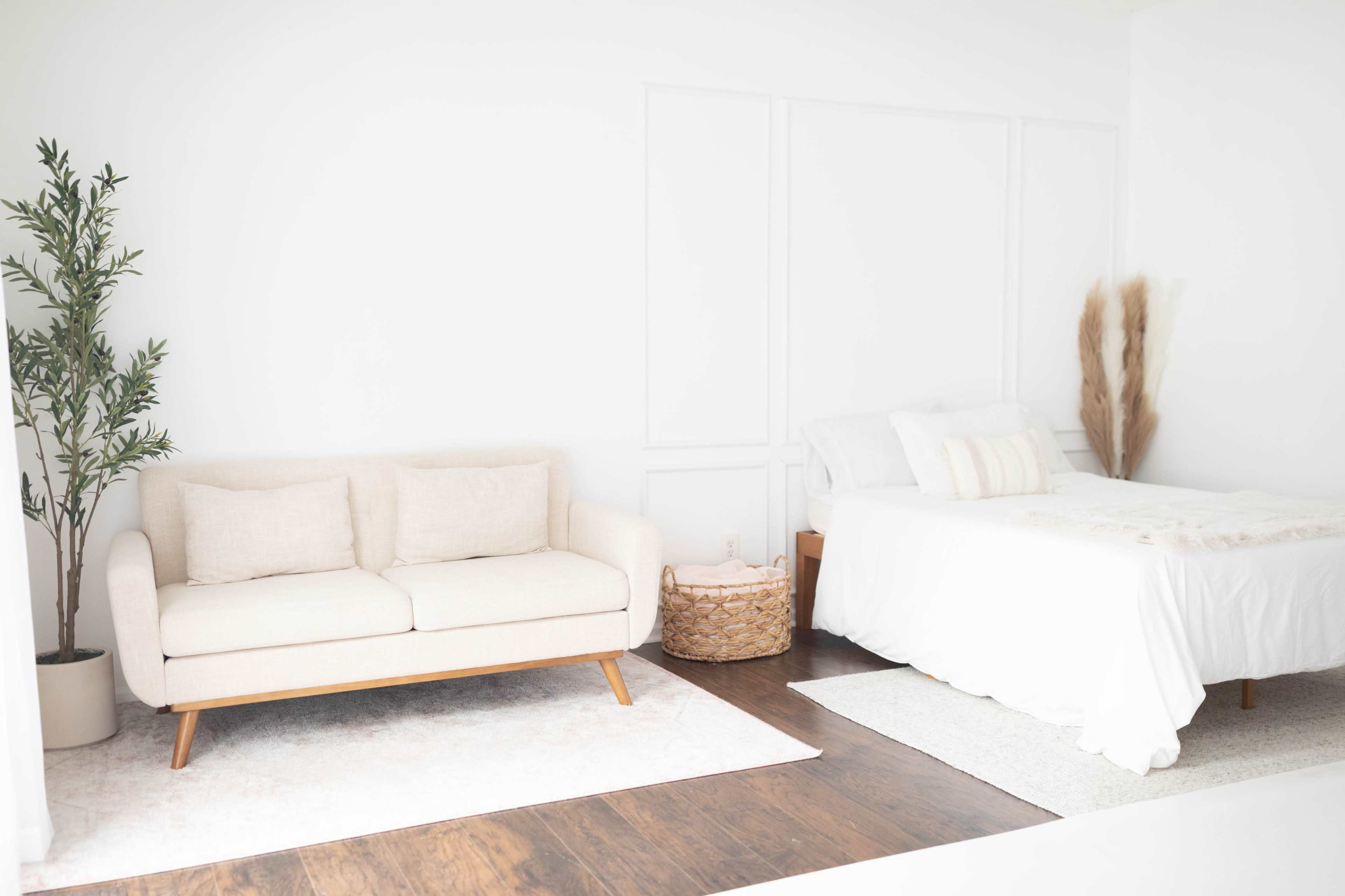 The image shows a bright, minimalistic bedroom featuring a bed, a light-colored sofa, and decorative plants.