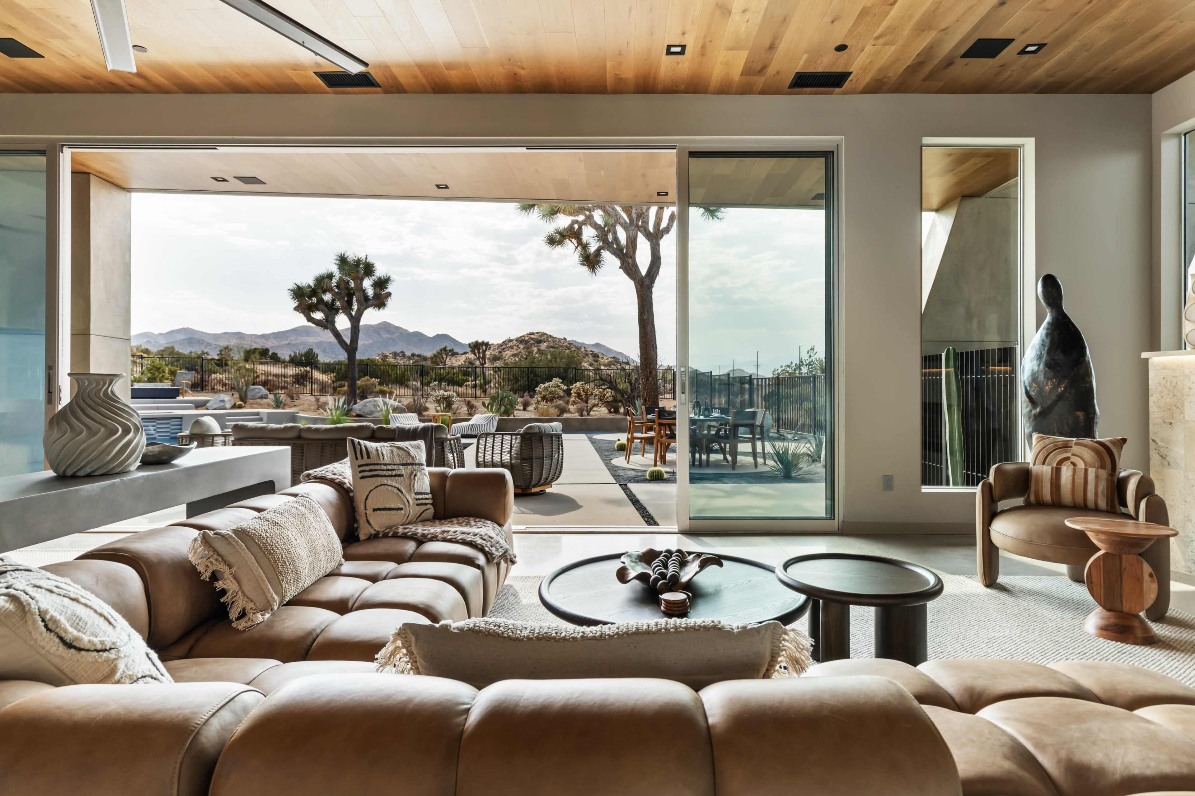 The image shows a modern living room with a large leather sofa, glass doors opening to a desert landscape, and a distinct sculpture in the corner.