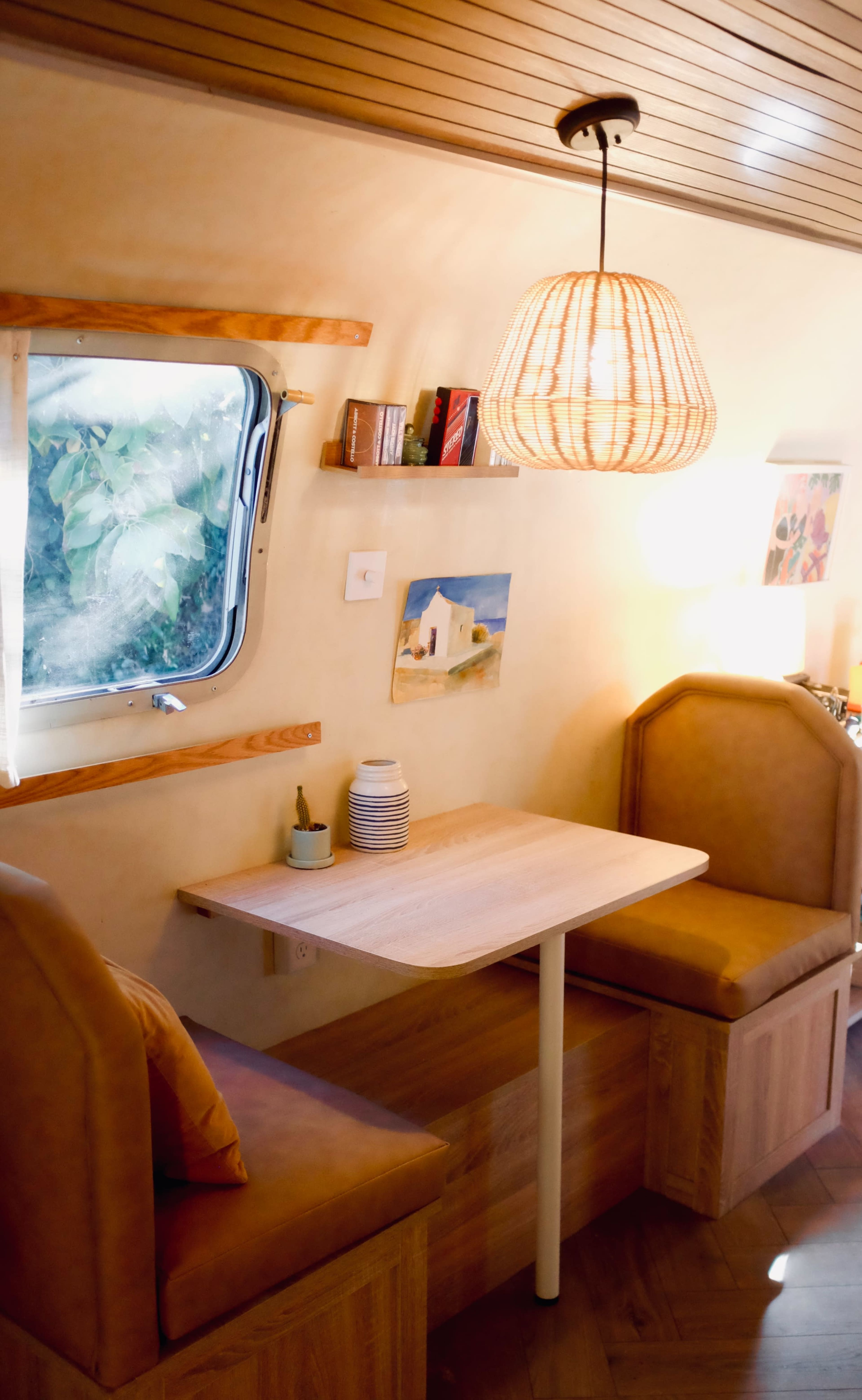 The image shows a cozy interior of a small living space with a table and two benches, a pendant light hanging from the ceiling, and a window letting in natural light.
