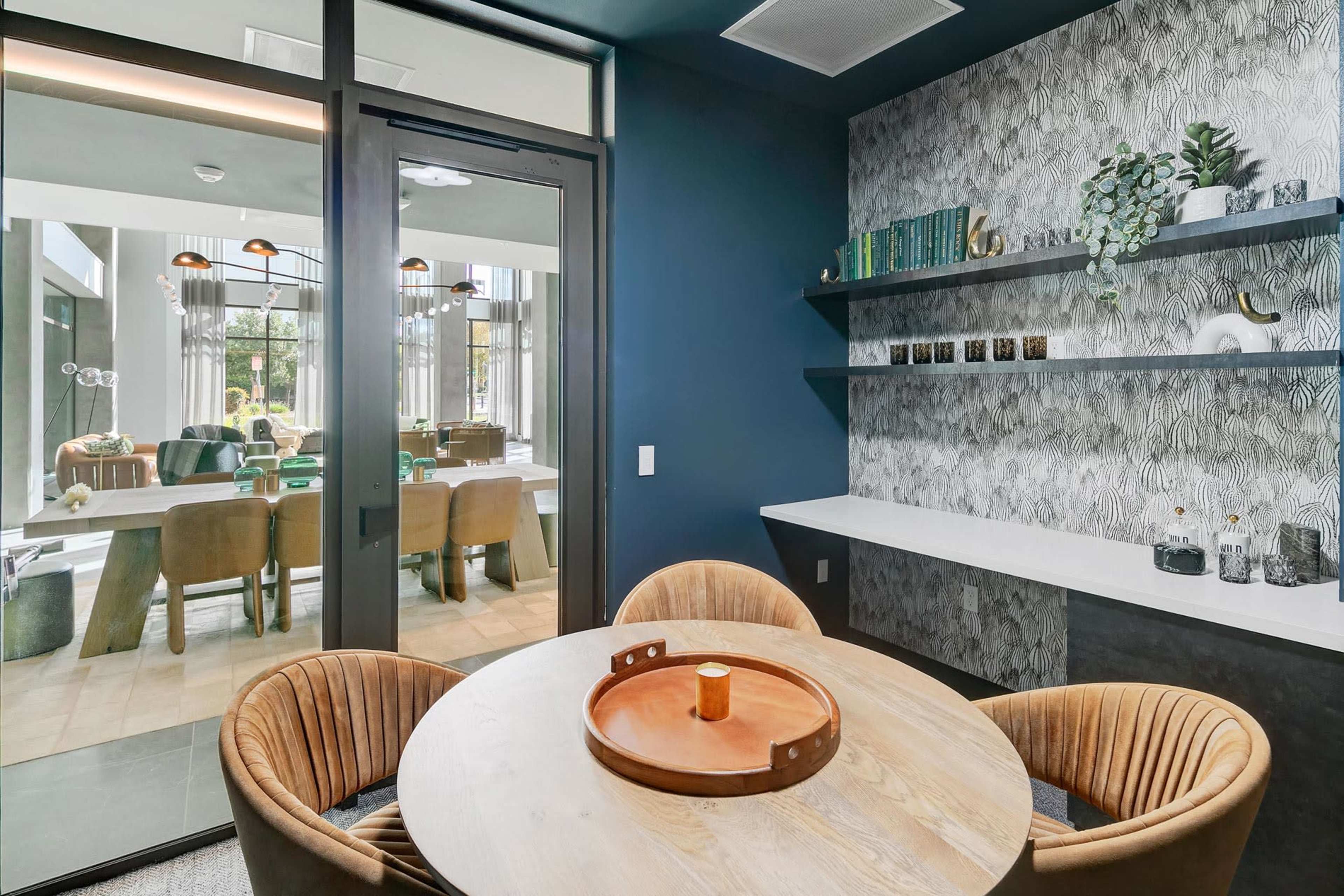 A modern dining area with round wooden chairs, a circular table, and a wall featuring patterned wallpaper and shelves displaying decorative items, with an open view into a well-lit room beyond.