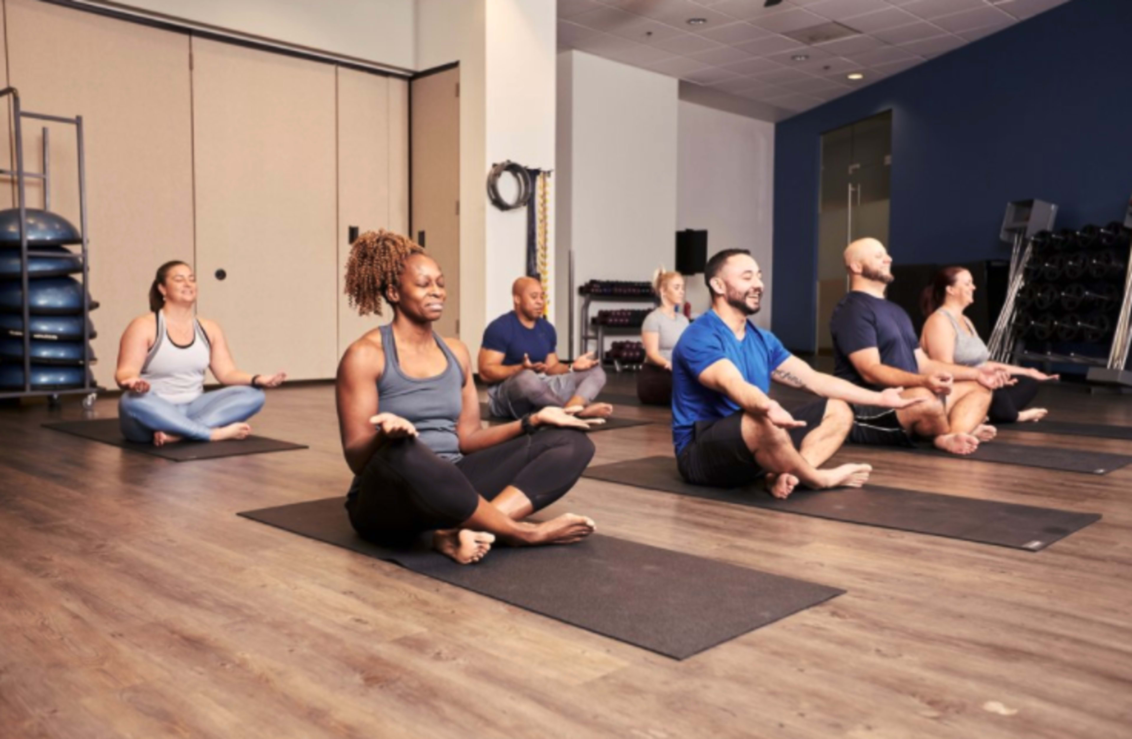 A group of people sits cross-legged on yoga mats in a fitness studio, practicing meditation.